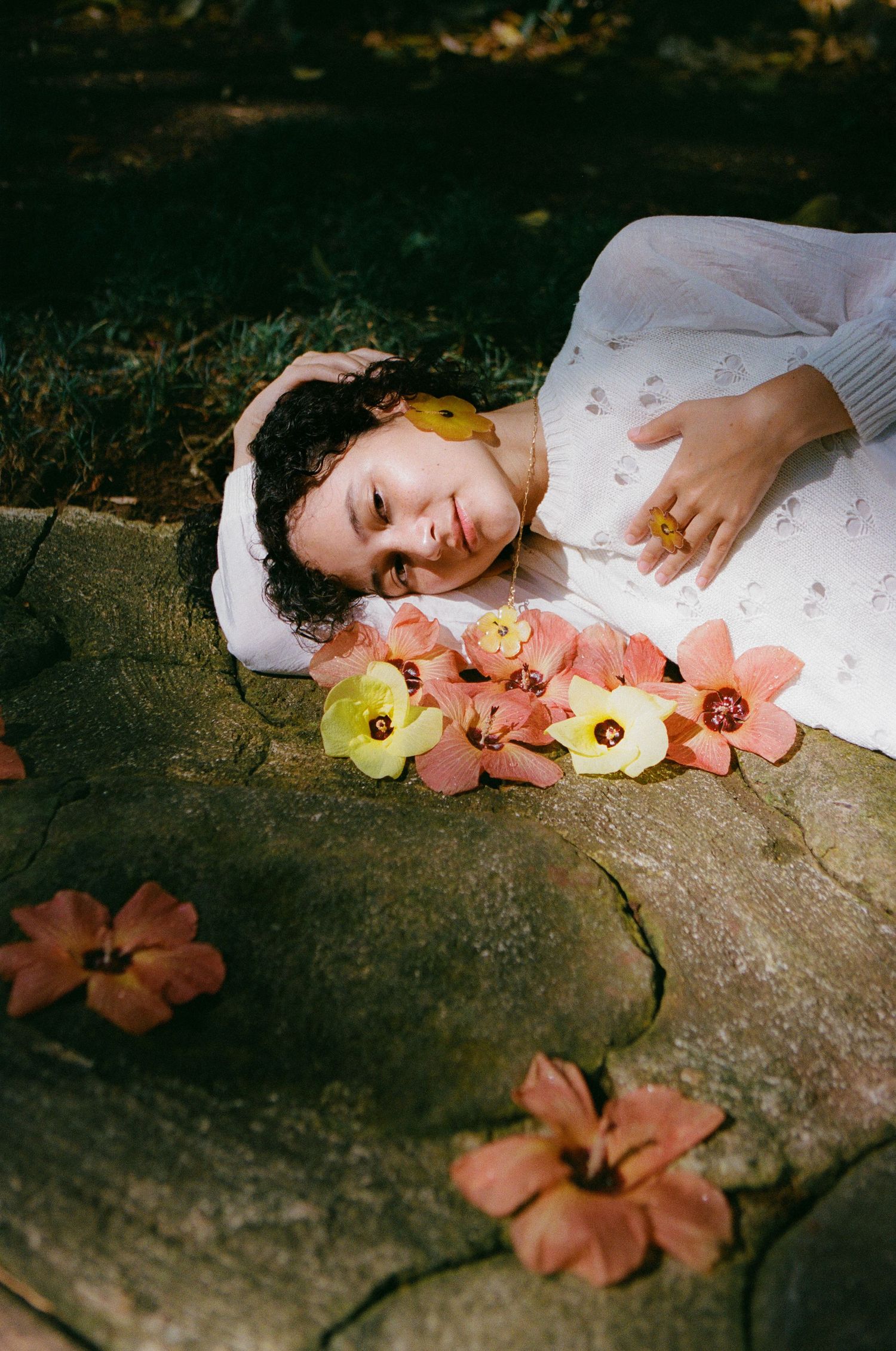 Woman in white dress lying on stone surrounded by pink and yellow tropical flowers.