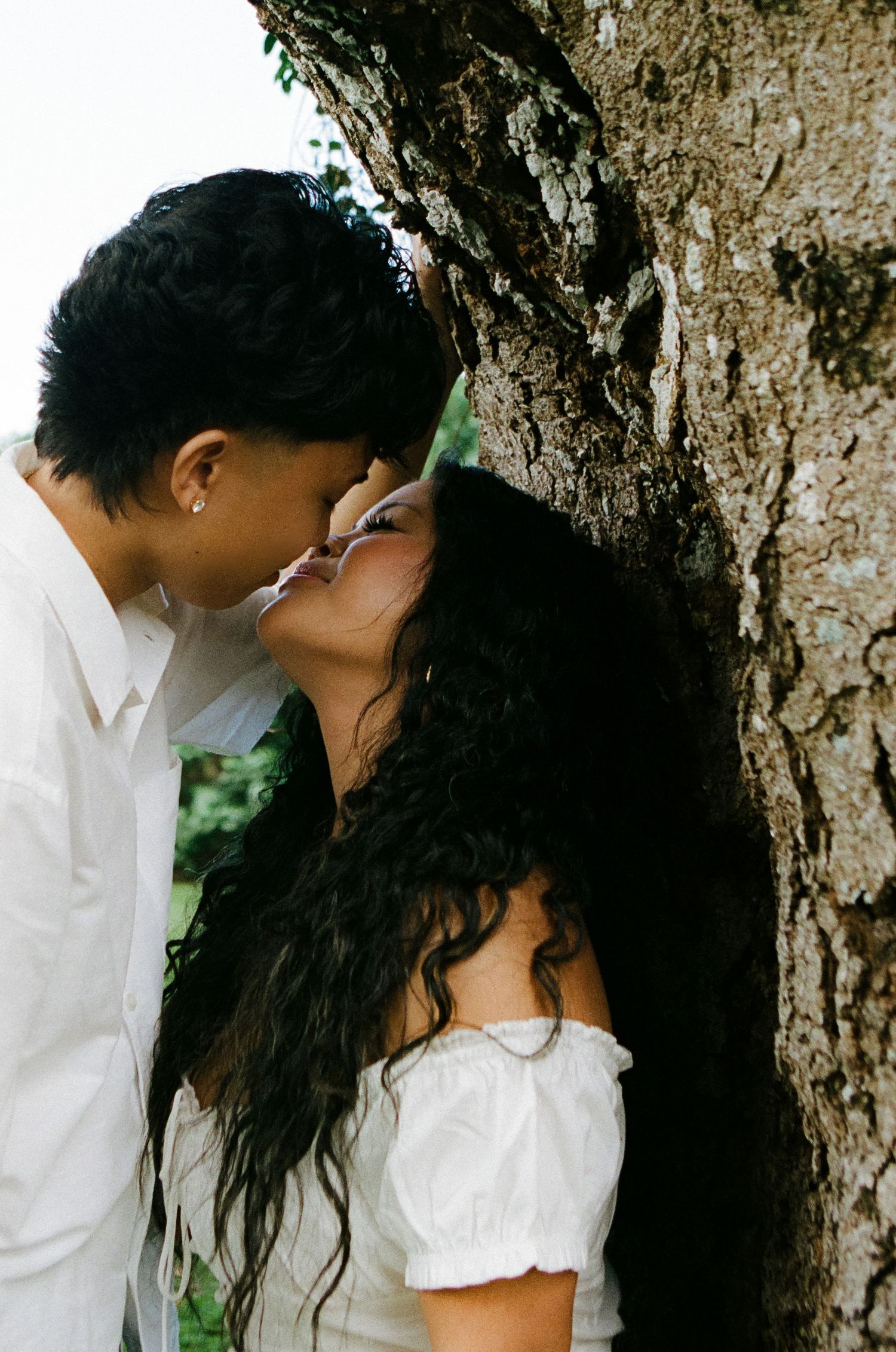 Couple shares a romantic kiss leaning against a large tree trunk in lush greenery.
