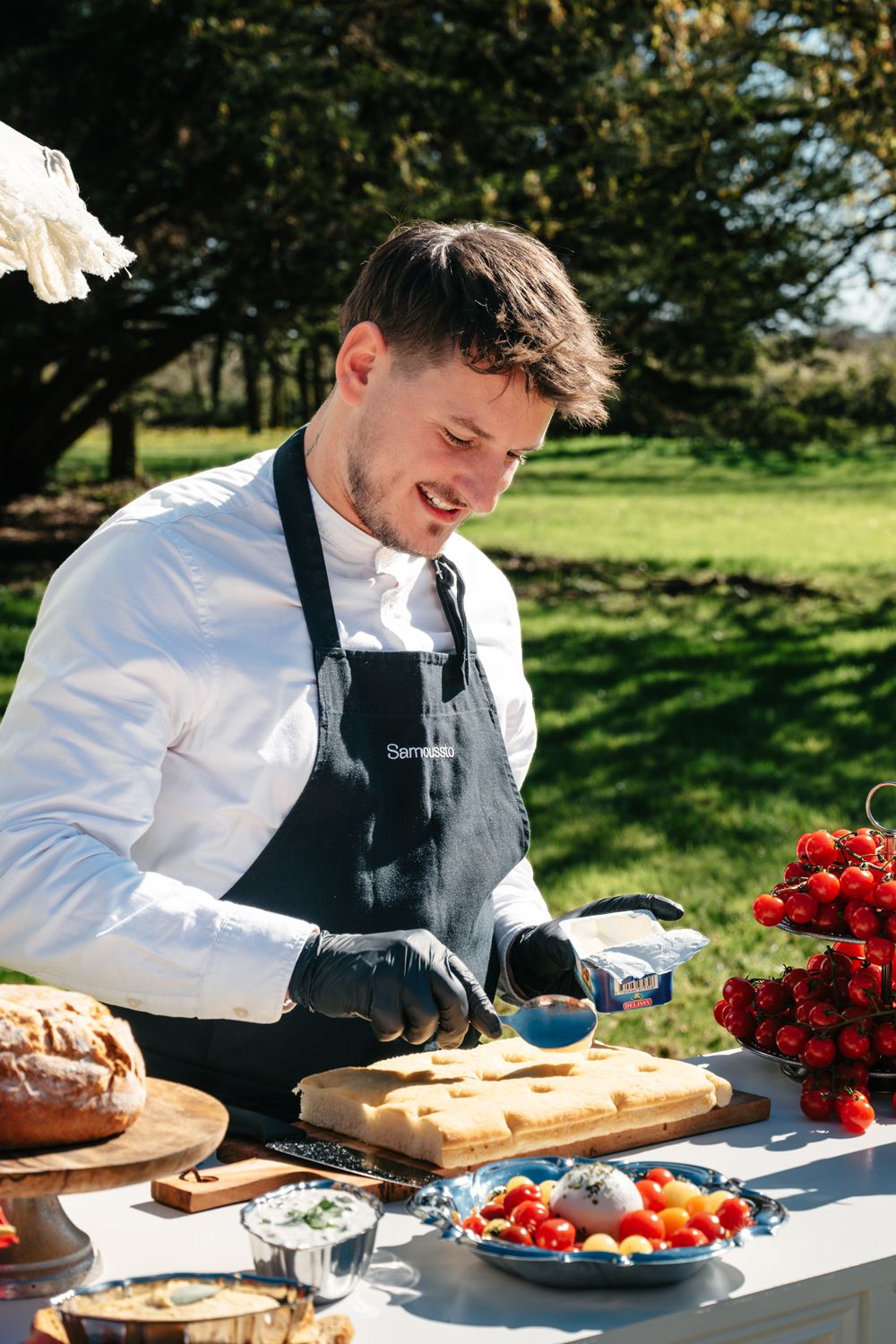 Un chef en tablier noir préparant un repas en plein air, sur une table rustique, avec des ingrédients frais, sur le thème Dolce Vita.