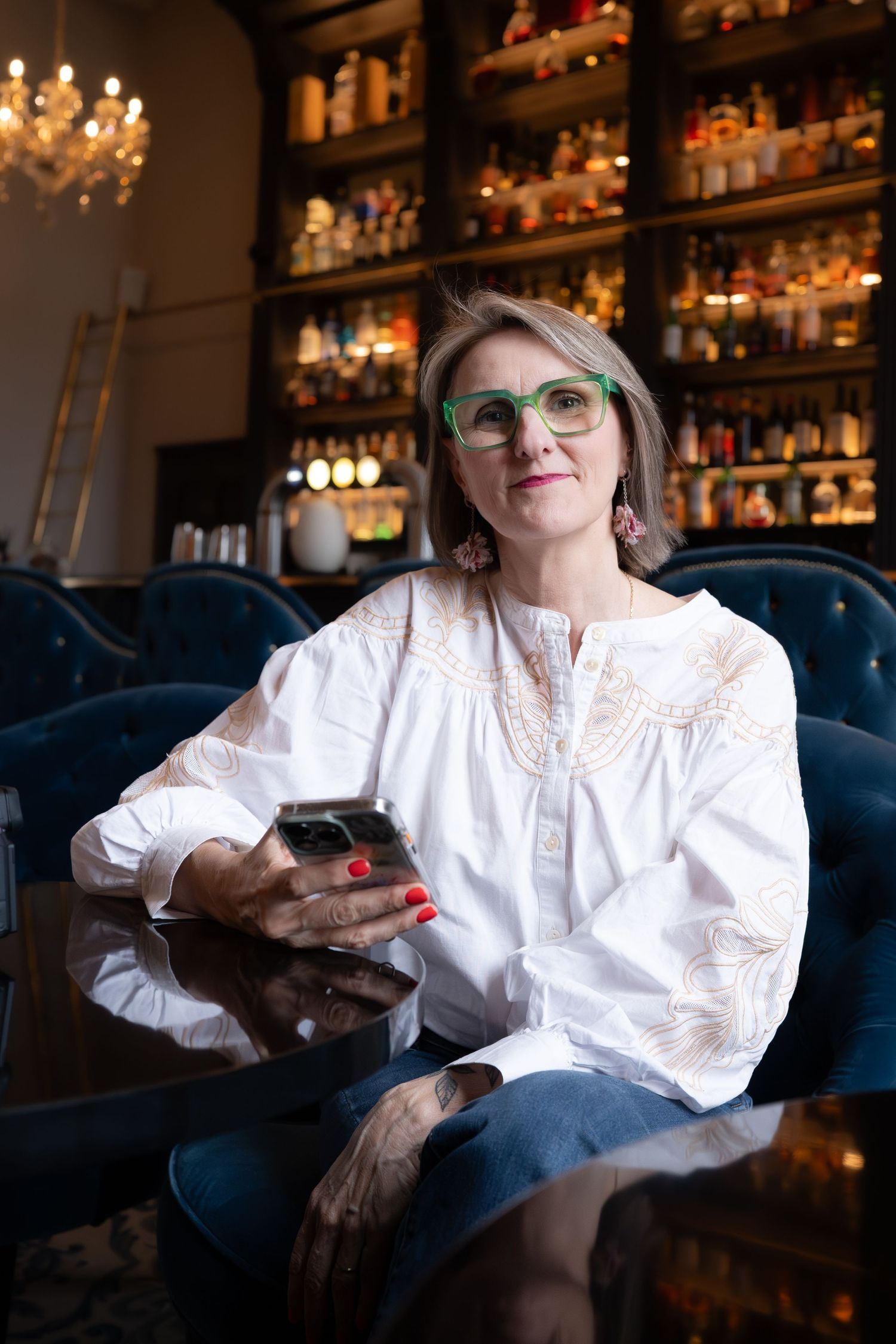 Une femme élégante, vêtue d'un chemisier blanc et portant des lunettes vertes, tenant un téléphone dans un décor de bar. Steph Lecaudé