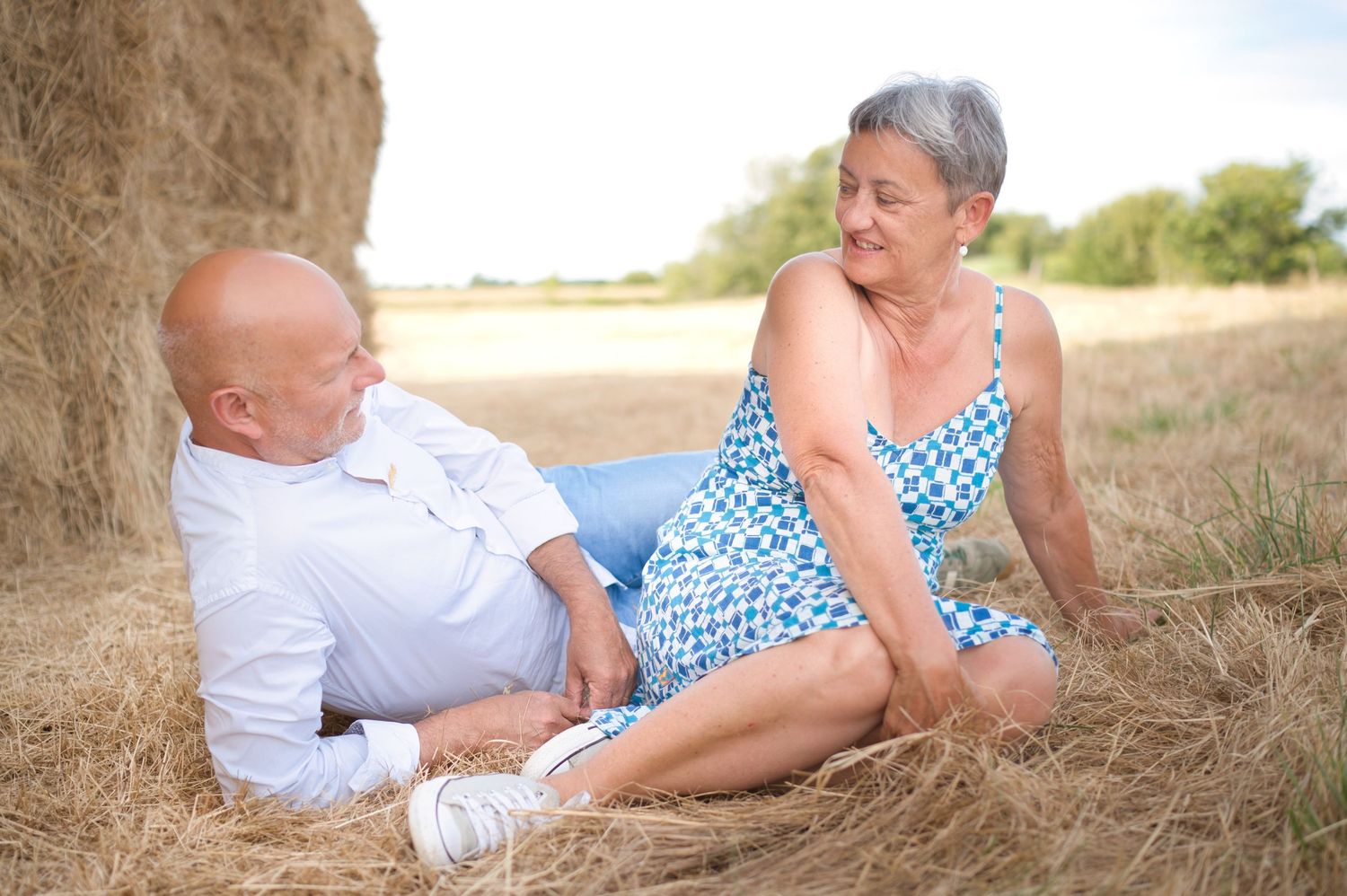 Un couple quinqua se détendant ensemble, adossés à une botte de foin dans un champ ensoleillé, souriant et se regardant.