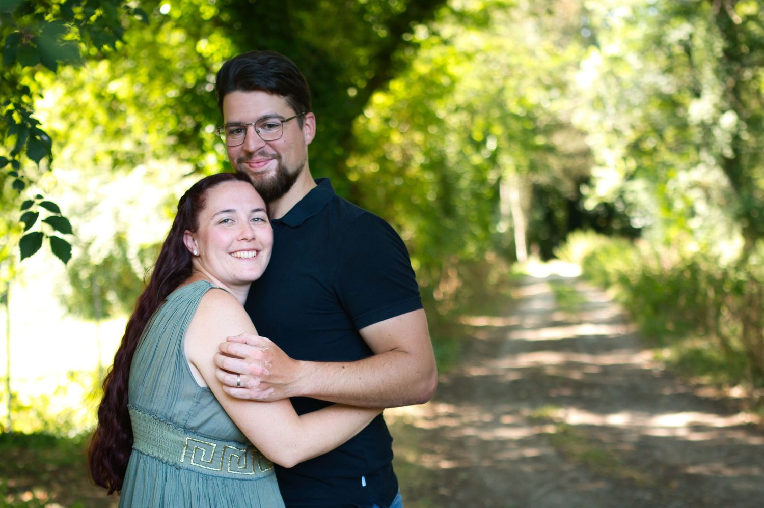 Un couple heureux s'étreignant sur un sentier bordé d'arbres ; la femme porte une robe couleur sauge et l'homme un polo noir.