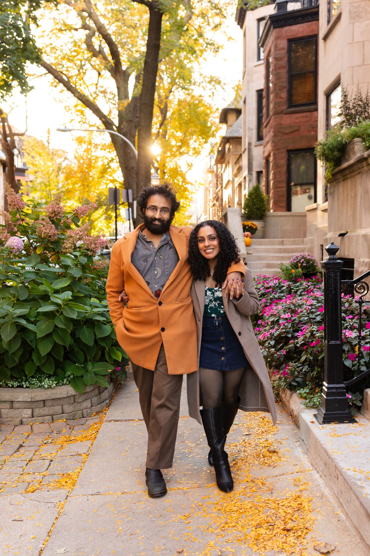 A smiling couple walks arm-in-arm along a flower-lined sidewalk during golden autumn sunset.
