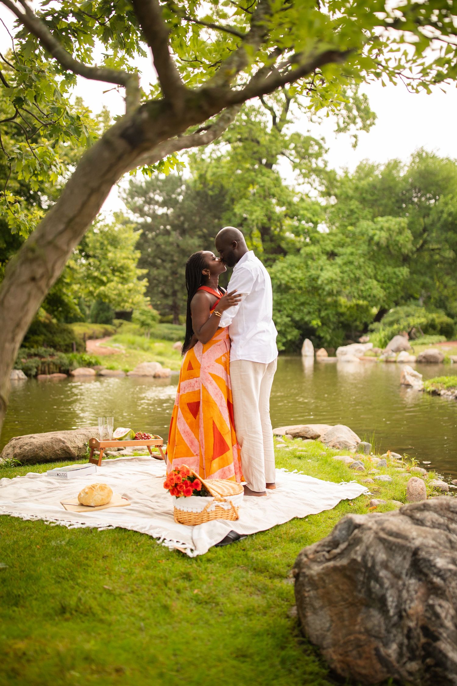 A couple embraces and kisses near a tranquil pond surrounded by lush greenery during a park picnic.