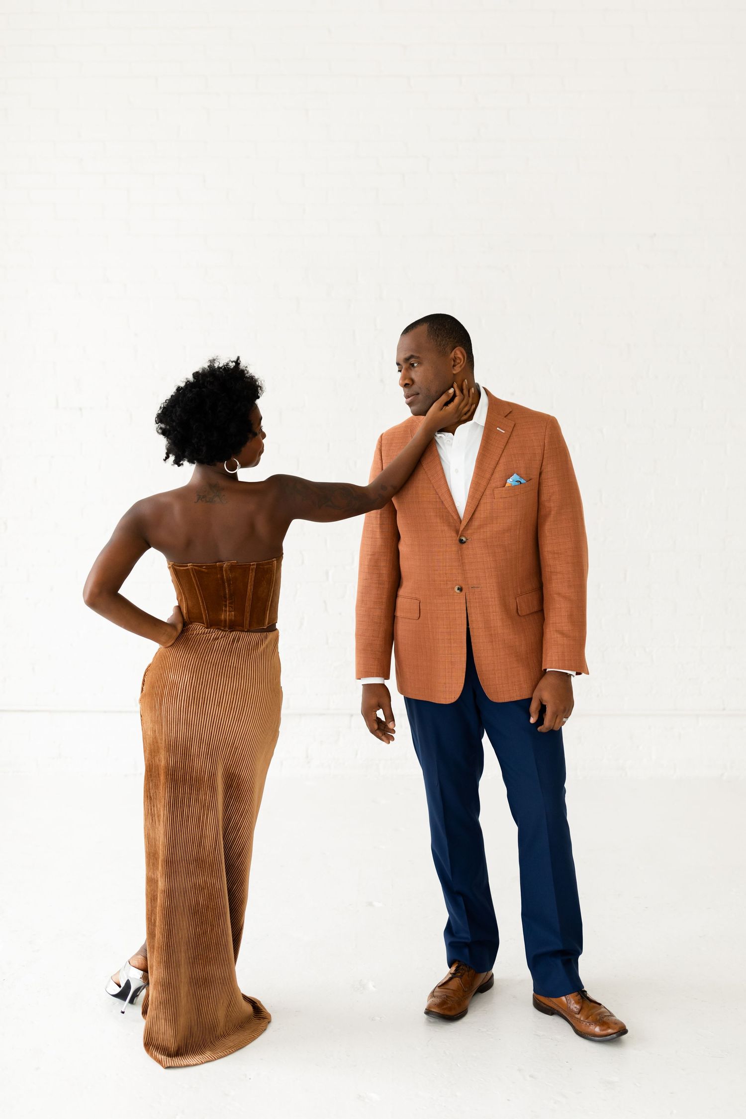 A couple poses elegantly, woman in brown velvet dress touches mans neck, he wears orange blazer and navy pants.