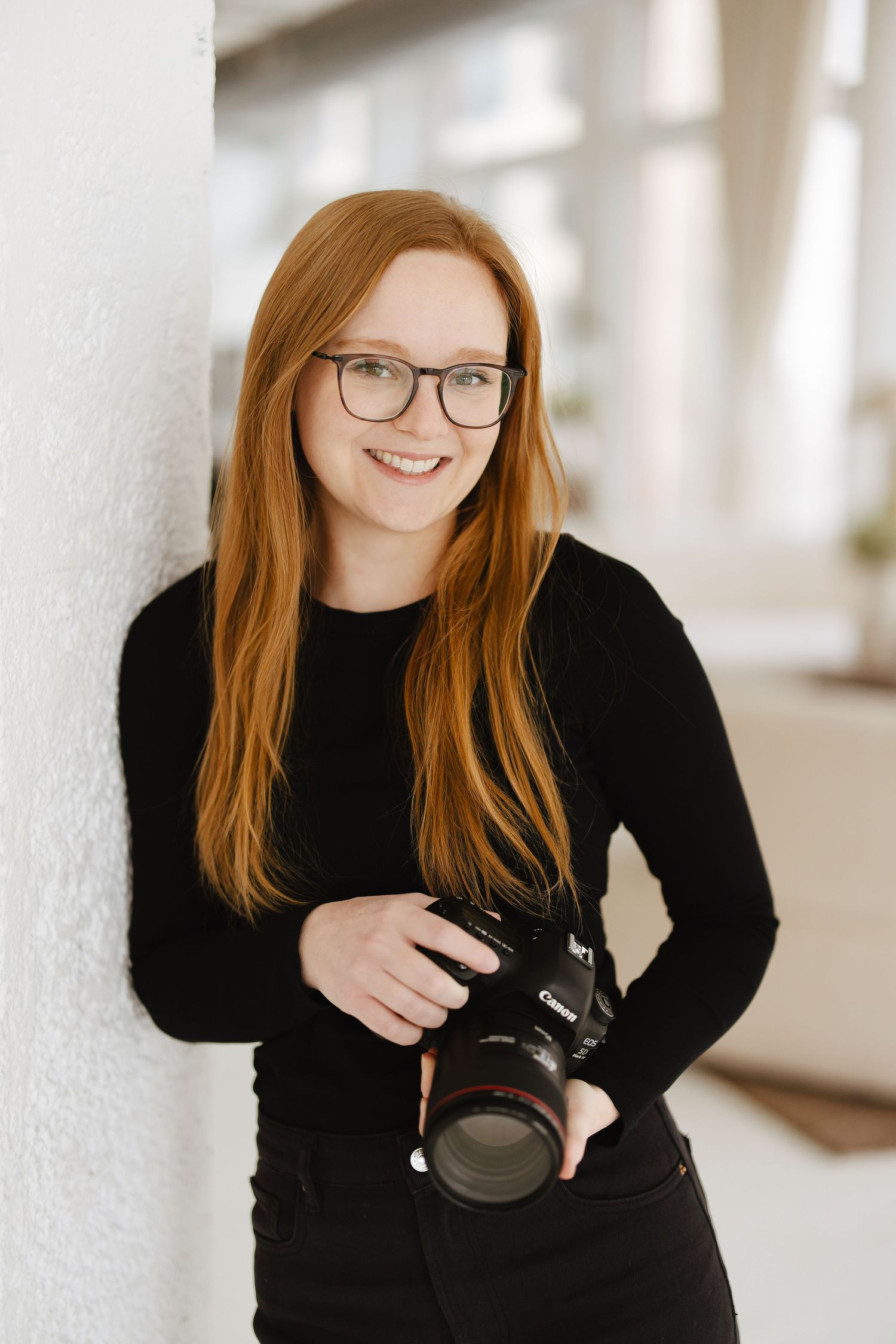 Young redhead female photographer with glasses holds Canon camera leaning against white wall.