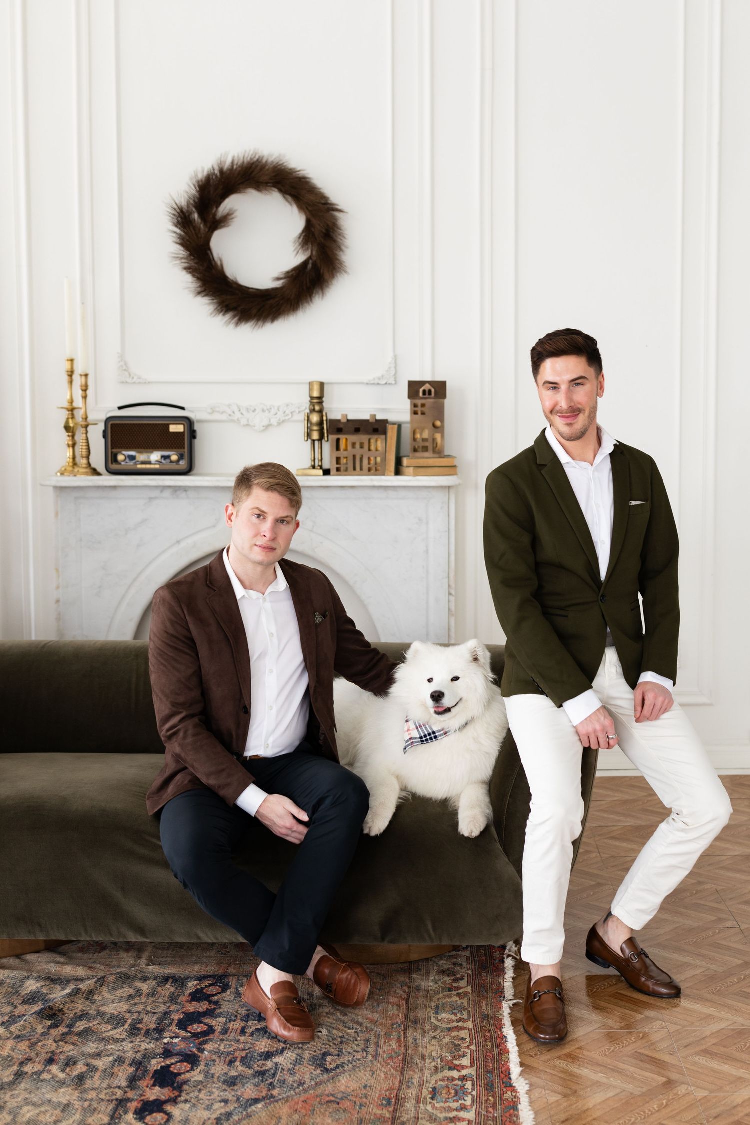 Two grooms in brown and green blazers sit with white Samoyed dog on green velvet sofa near fireplace.