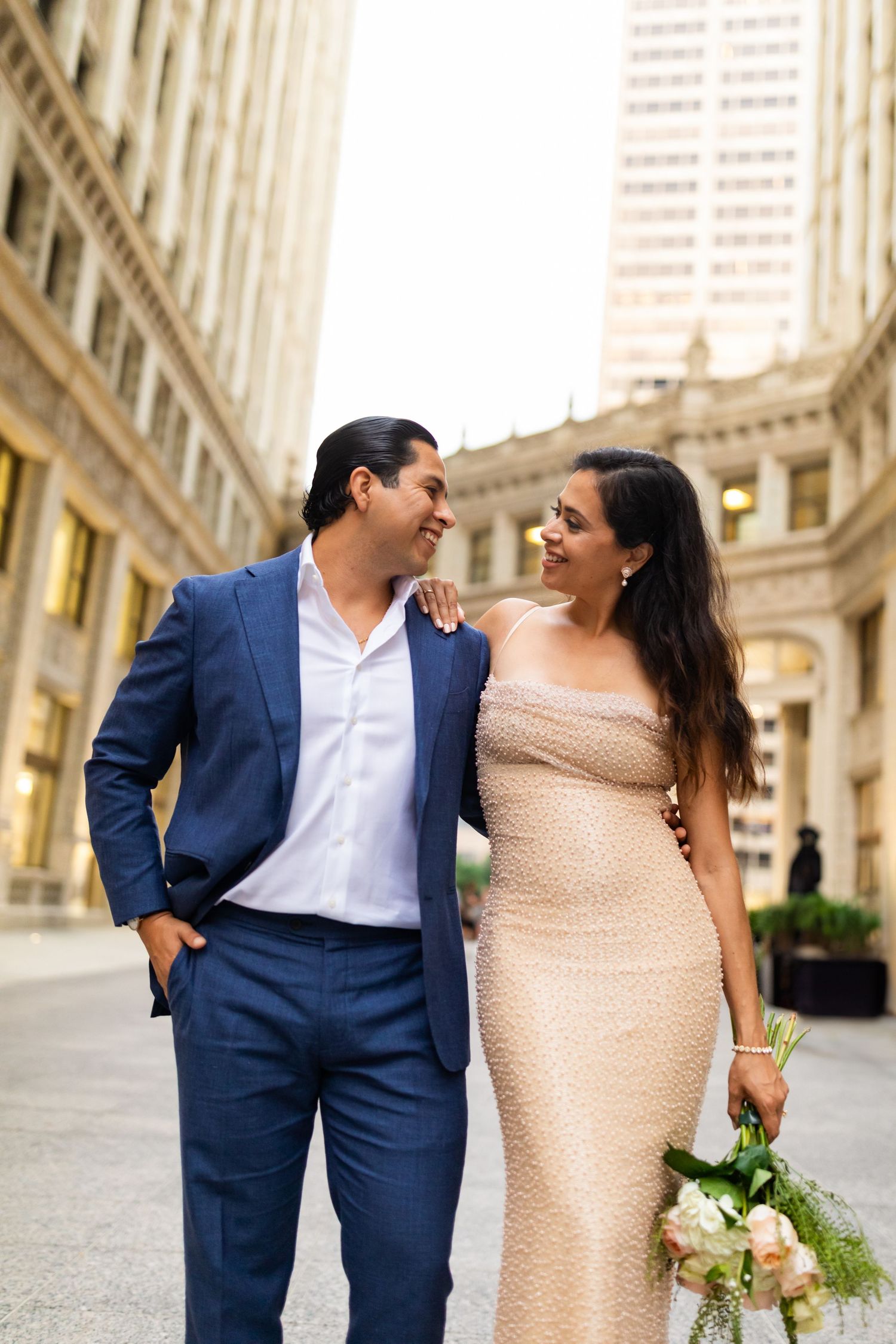 Smiling couple in formal wear walking together through Chicago urban corridor, woman holding white bouquet.