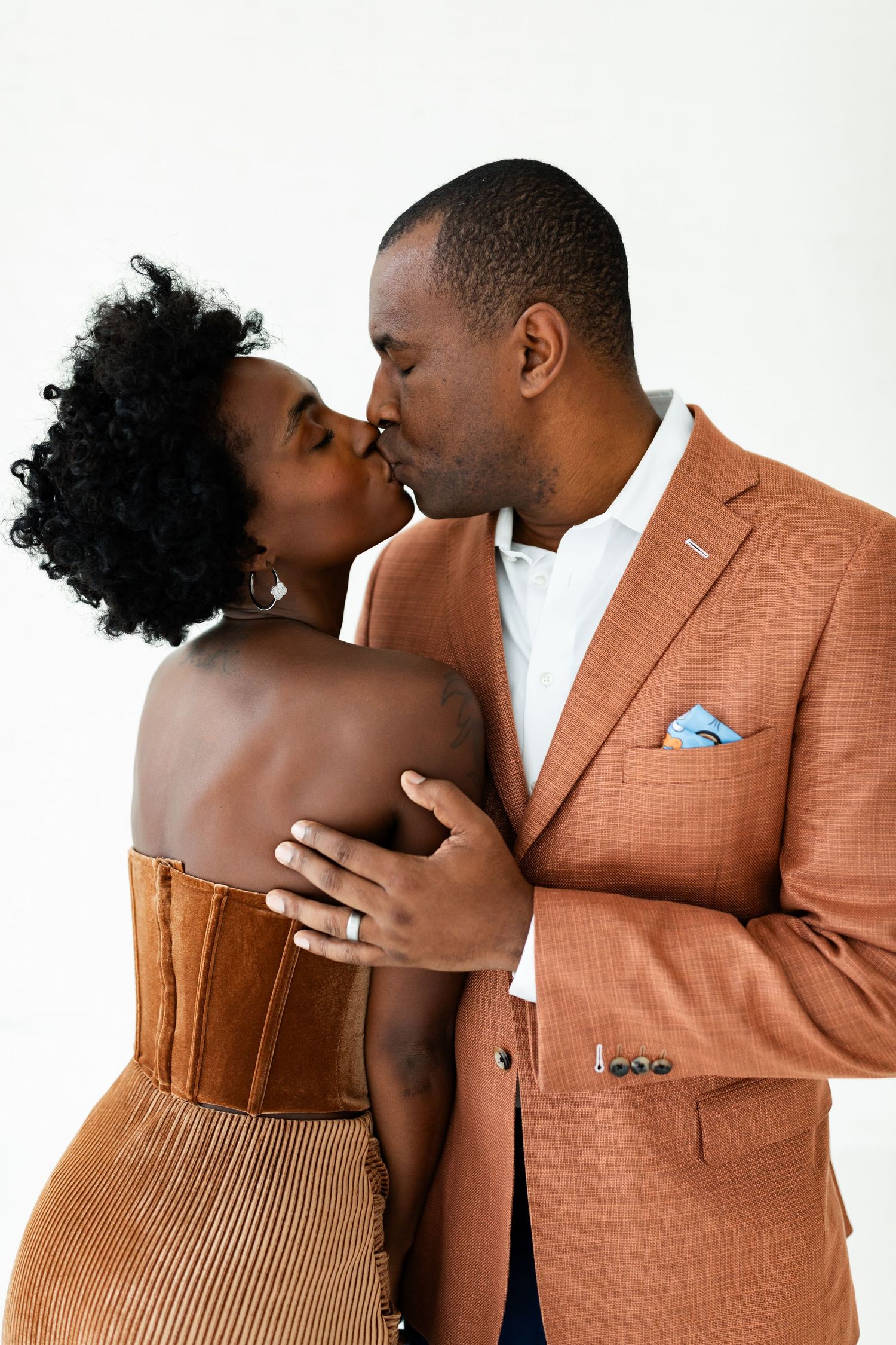 Romantic couple sharing a kiss, woman in brown velvet dress, man in orange plaid blazer with blue pocket square.