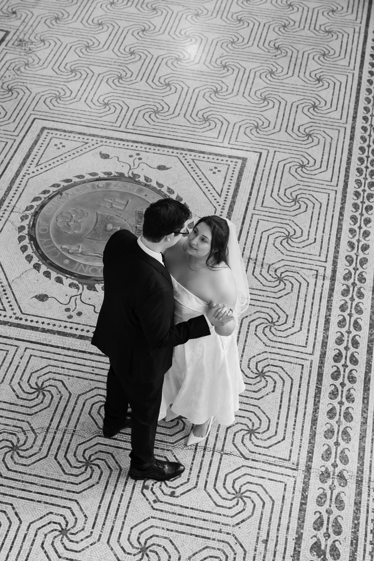 Black and white aerial view of a bride and groom embracing on an ornate mosaic tile floor.