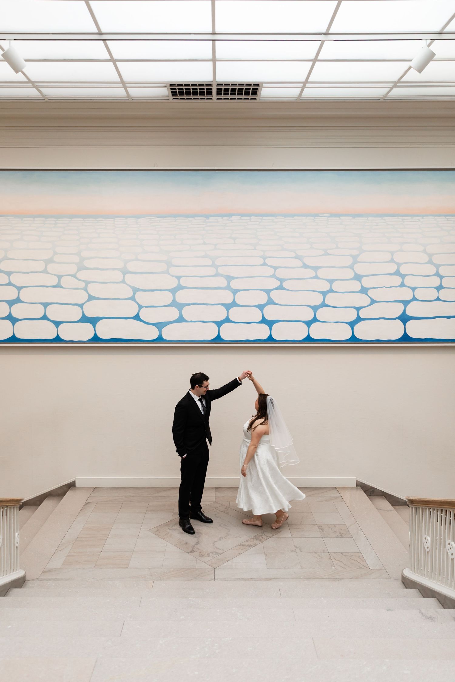 Groom spinning bride in a graceful dance beneath a large modern painting in a bright museum hall.