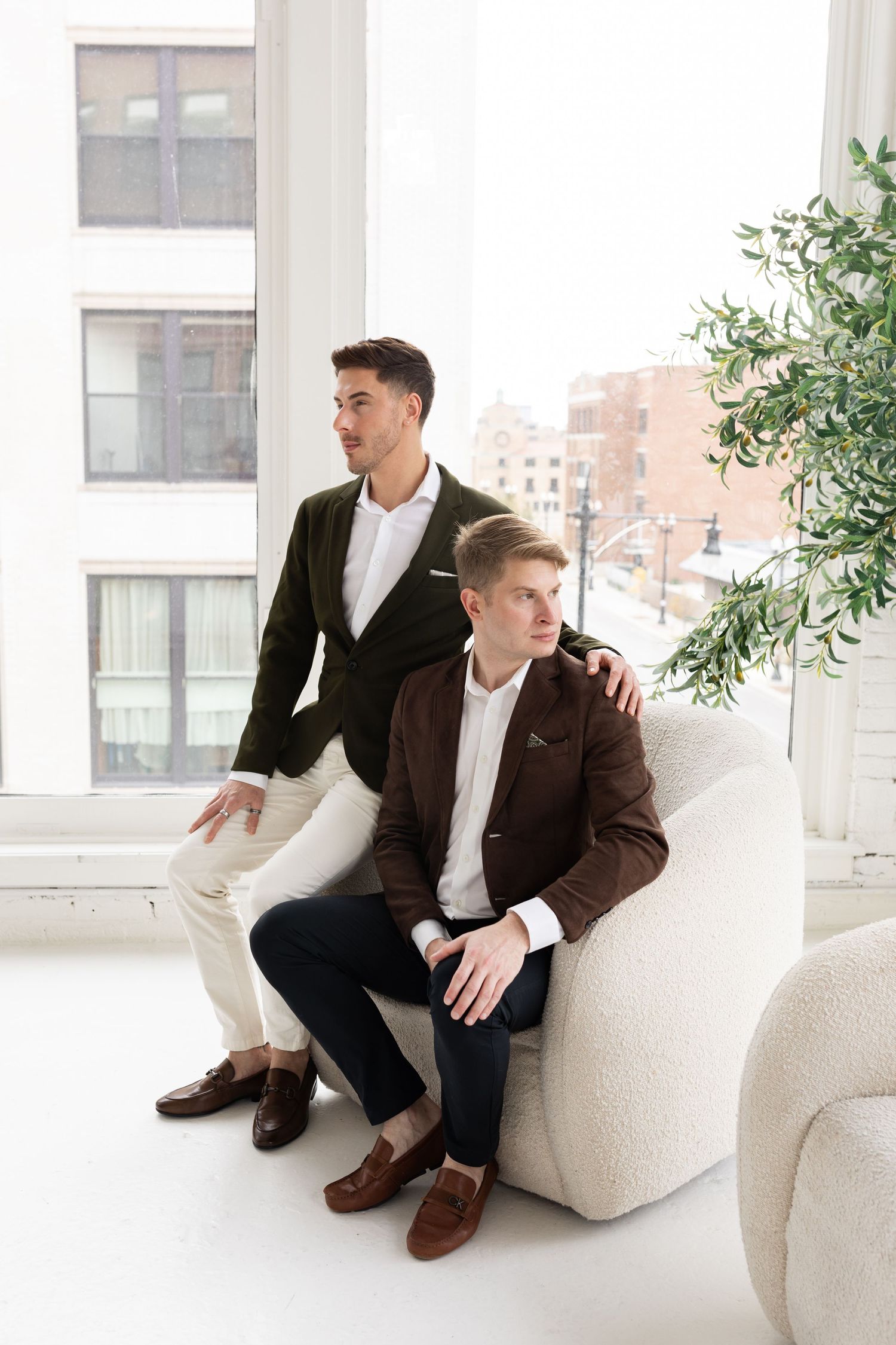 Two grooms in coordinating blazers pose near bright windows, one standing behind seated partner in white chair.