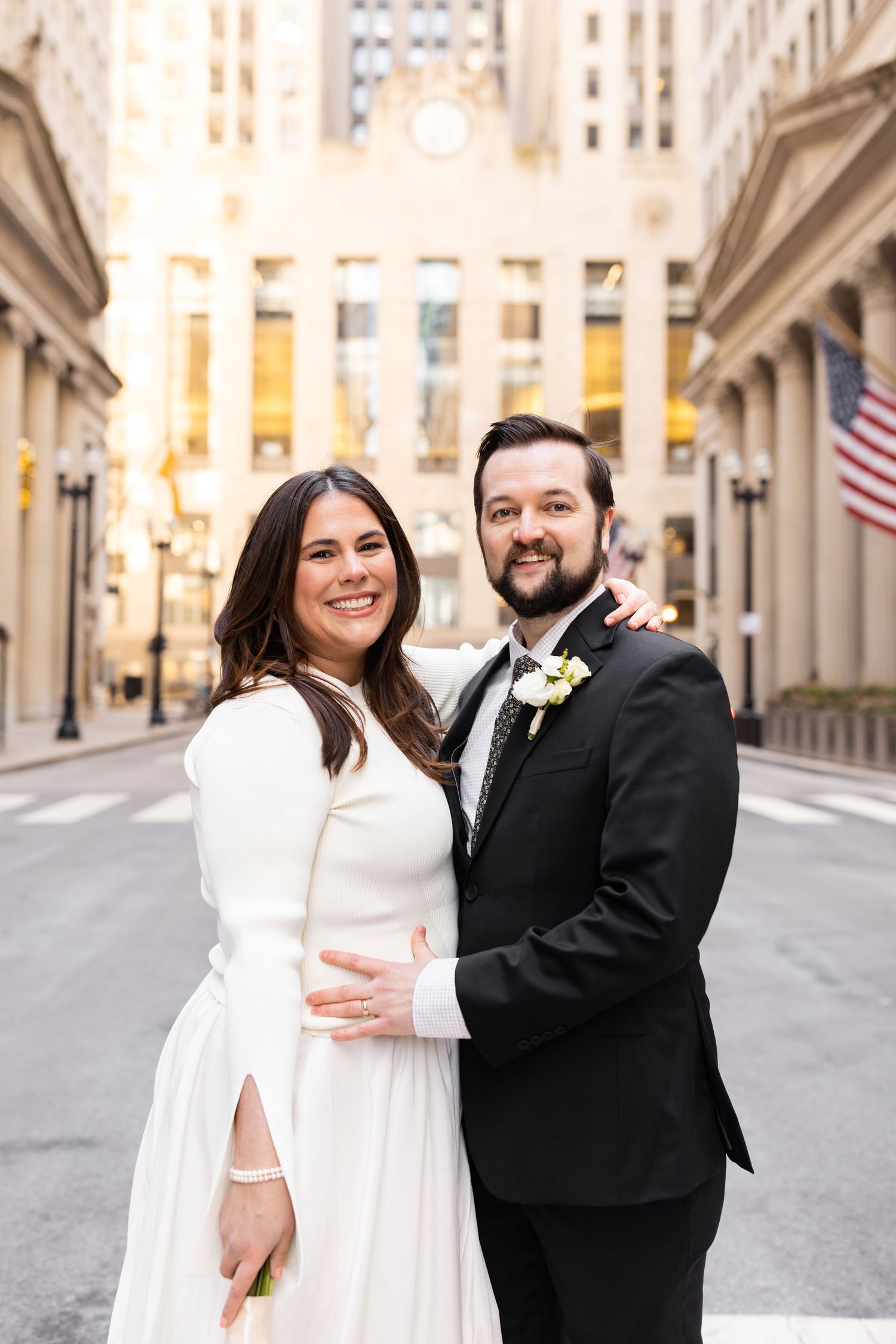Smiling newlyweds pose together on a city street, groom in black suit and bride in white cape dress.