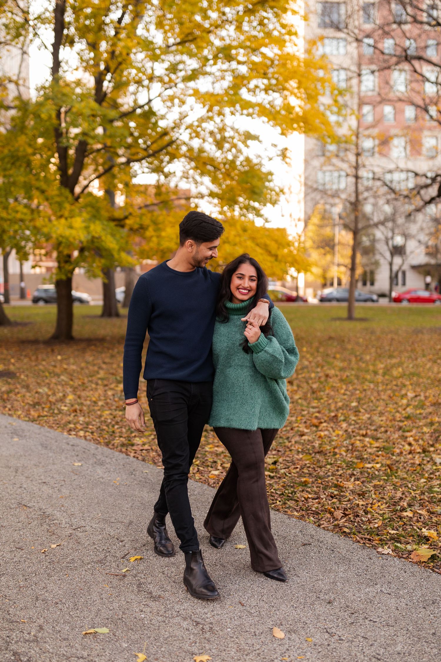 Couple walks arm in arm along a path in a park surrounded by vibrant autumn foliage.