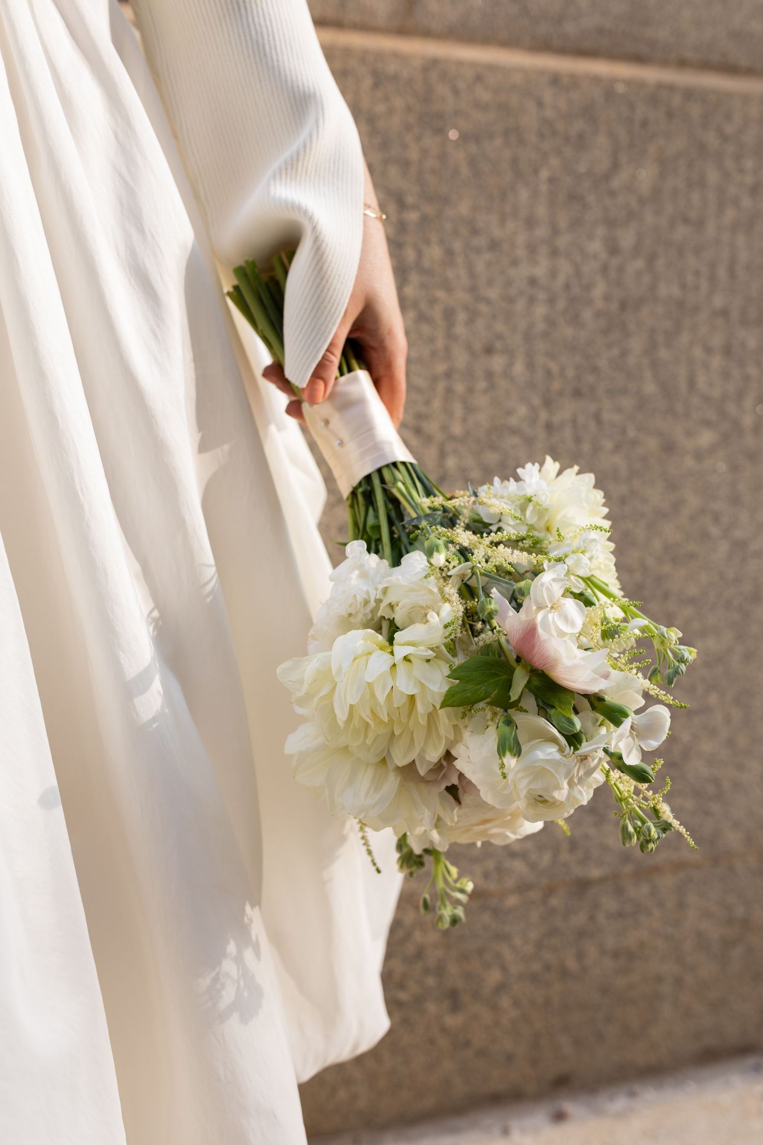 Bride holds elegant white and blush wedding bouquet with peonies and greenery against stone wall.