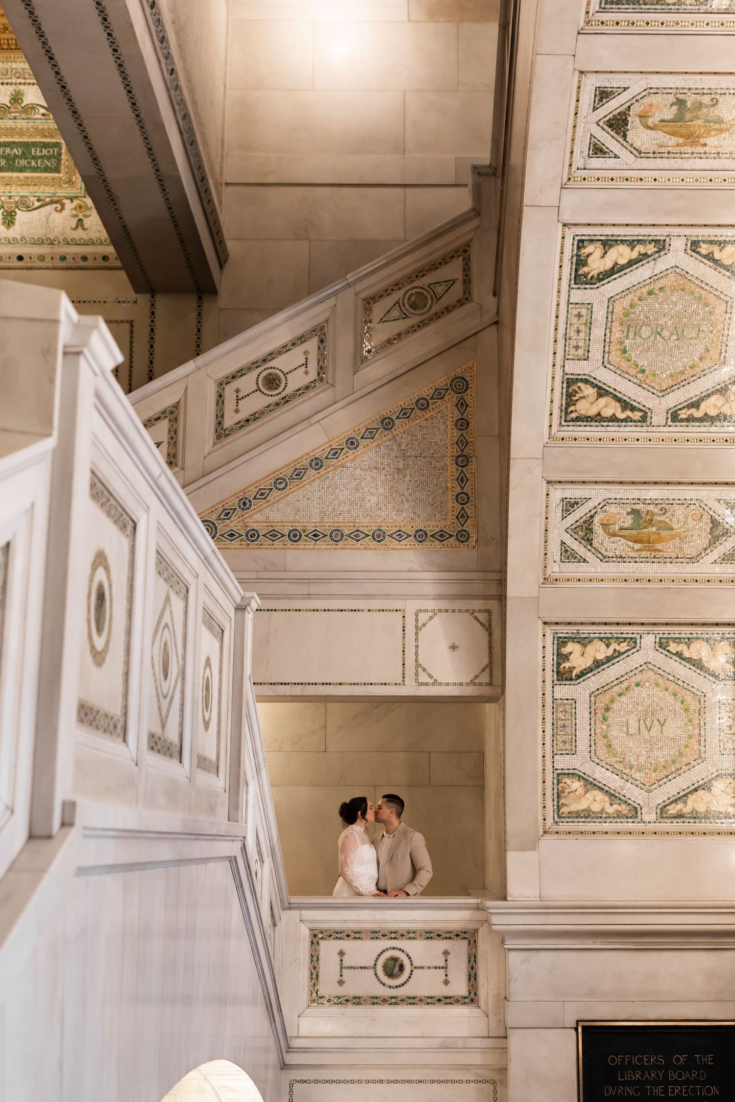 Bride and groom sharing a kiss on a balcony inside a grand ornate building with mosaic walls.