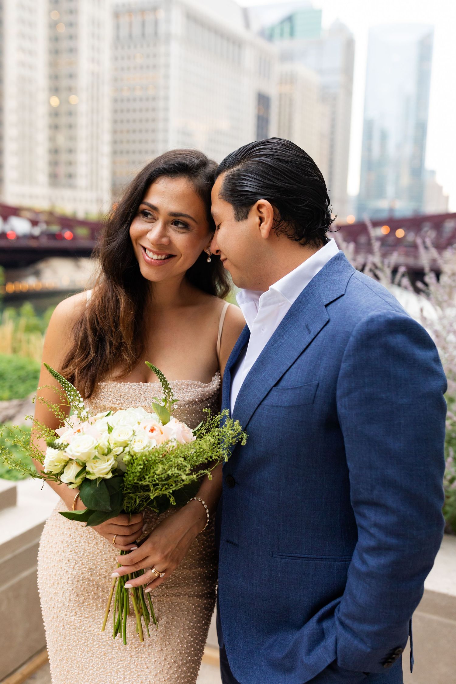 Bride smiling holding white roses while groom whispers to her on Chicago Riverwalk terrace.