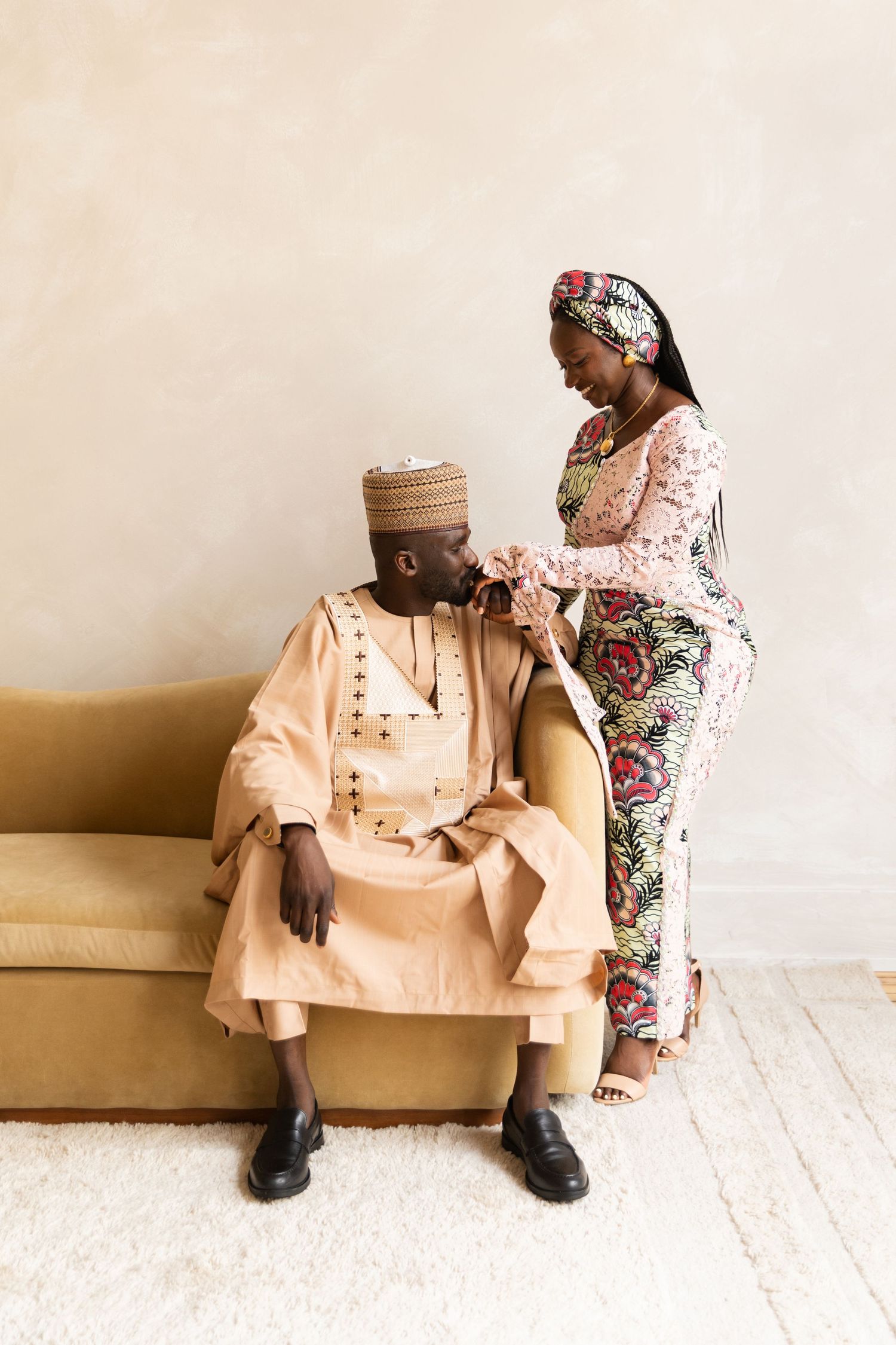 Couple in traditional Nigerian attire, man seated on sofa as woman tenderly holds his hands.