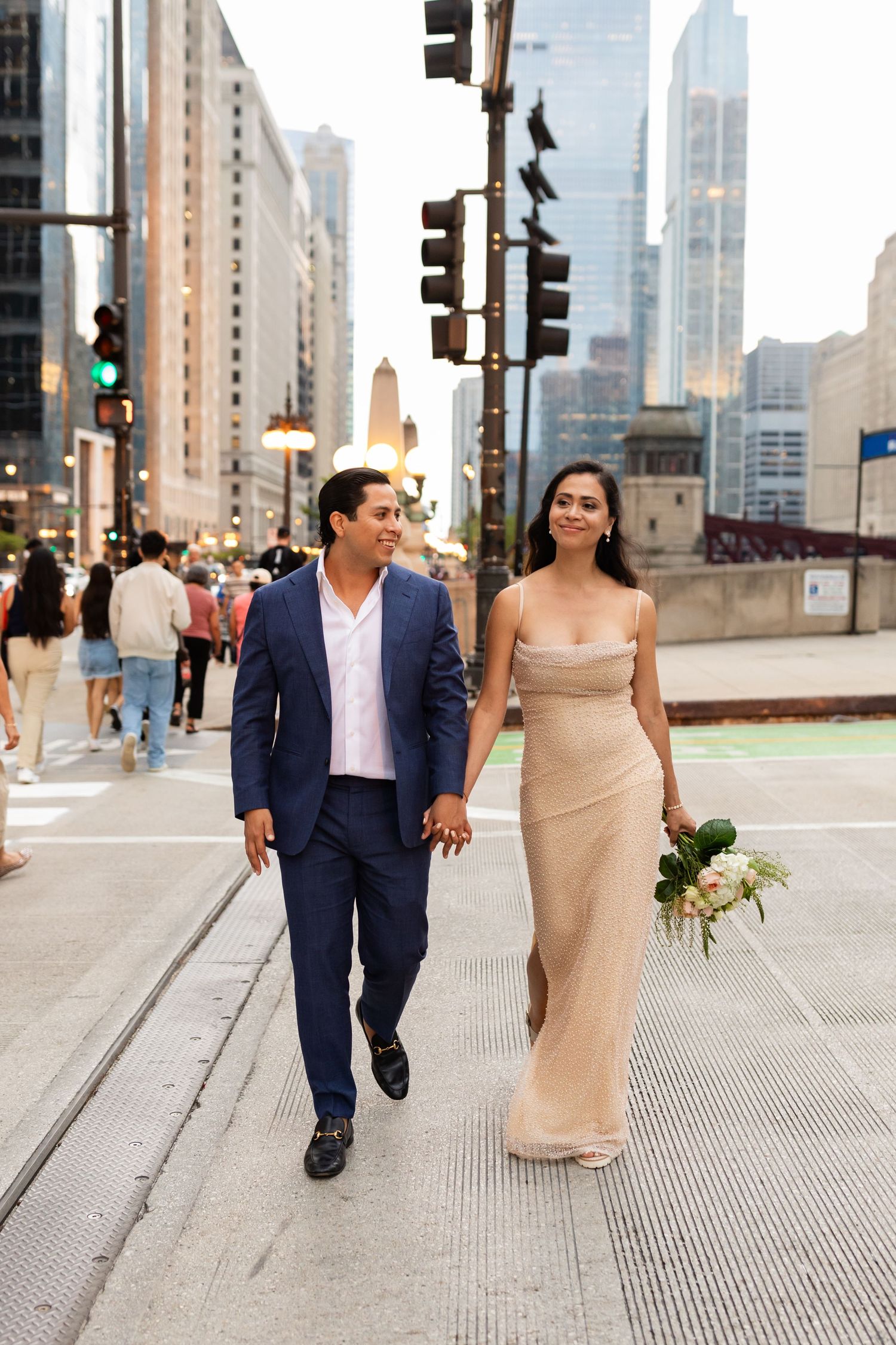 Newlywed couple walking hand in hand through Chicago streets at dusk, bride carrying white bouquet.