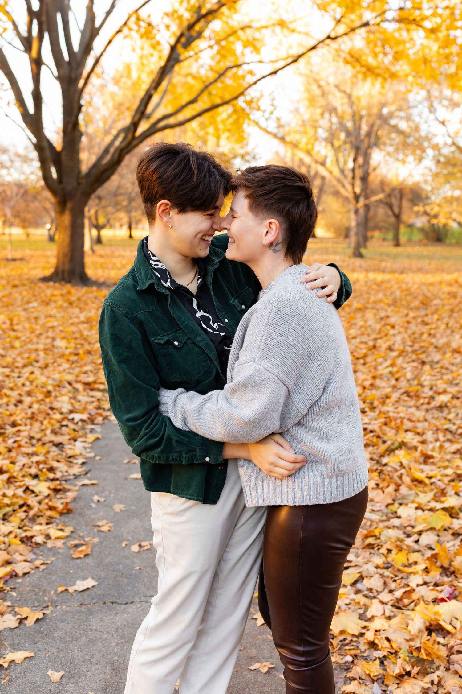 Two women embrace and smile at each other in an autumn park surrounded by golden fallen leaves.