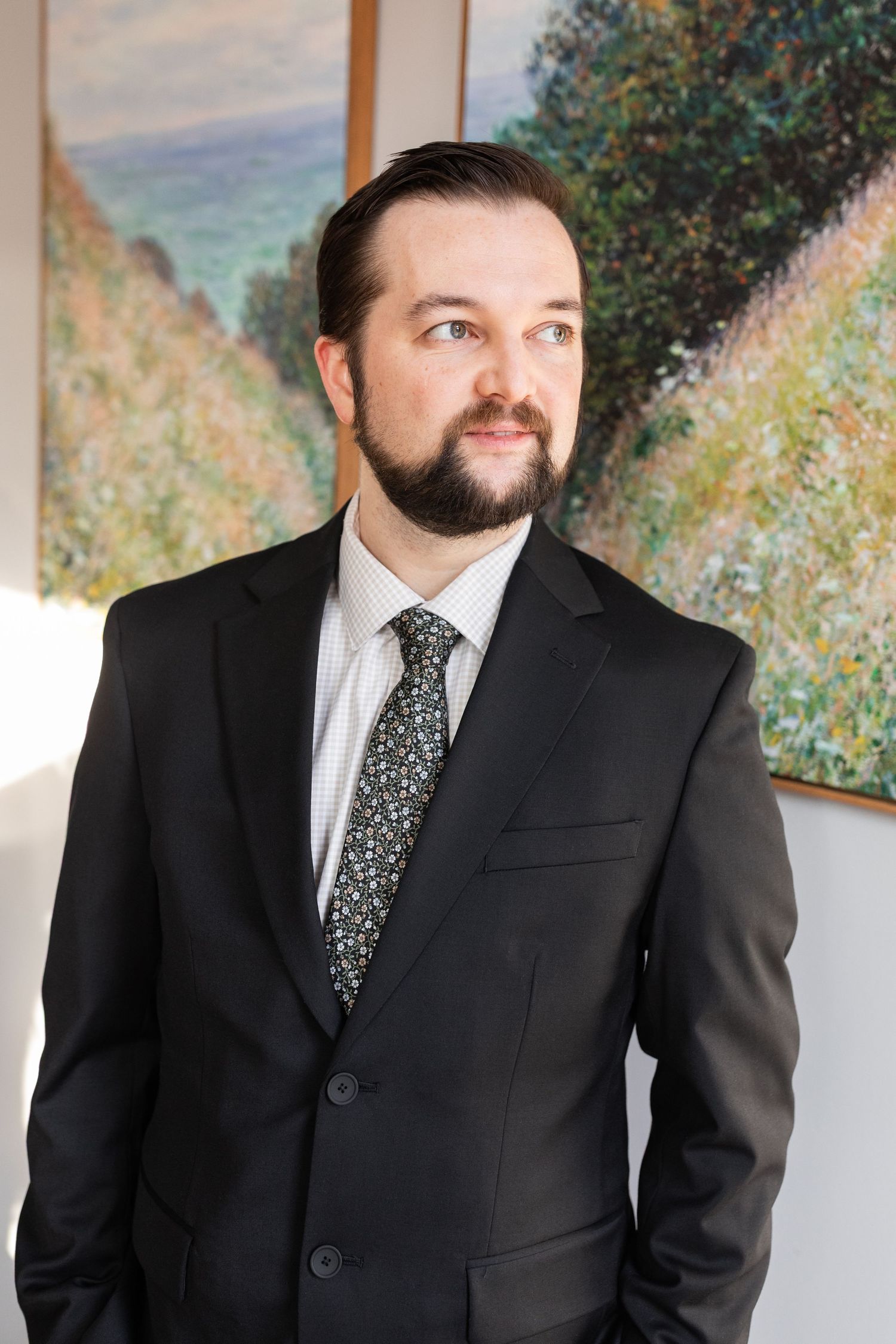 Bearded groom in black suit and patterned tie gazes sideways near impressionist artwork.