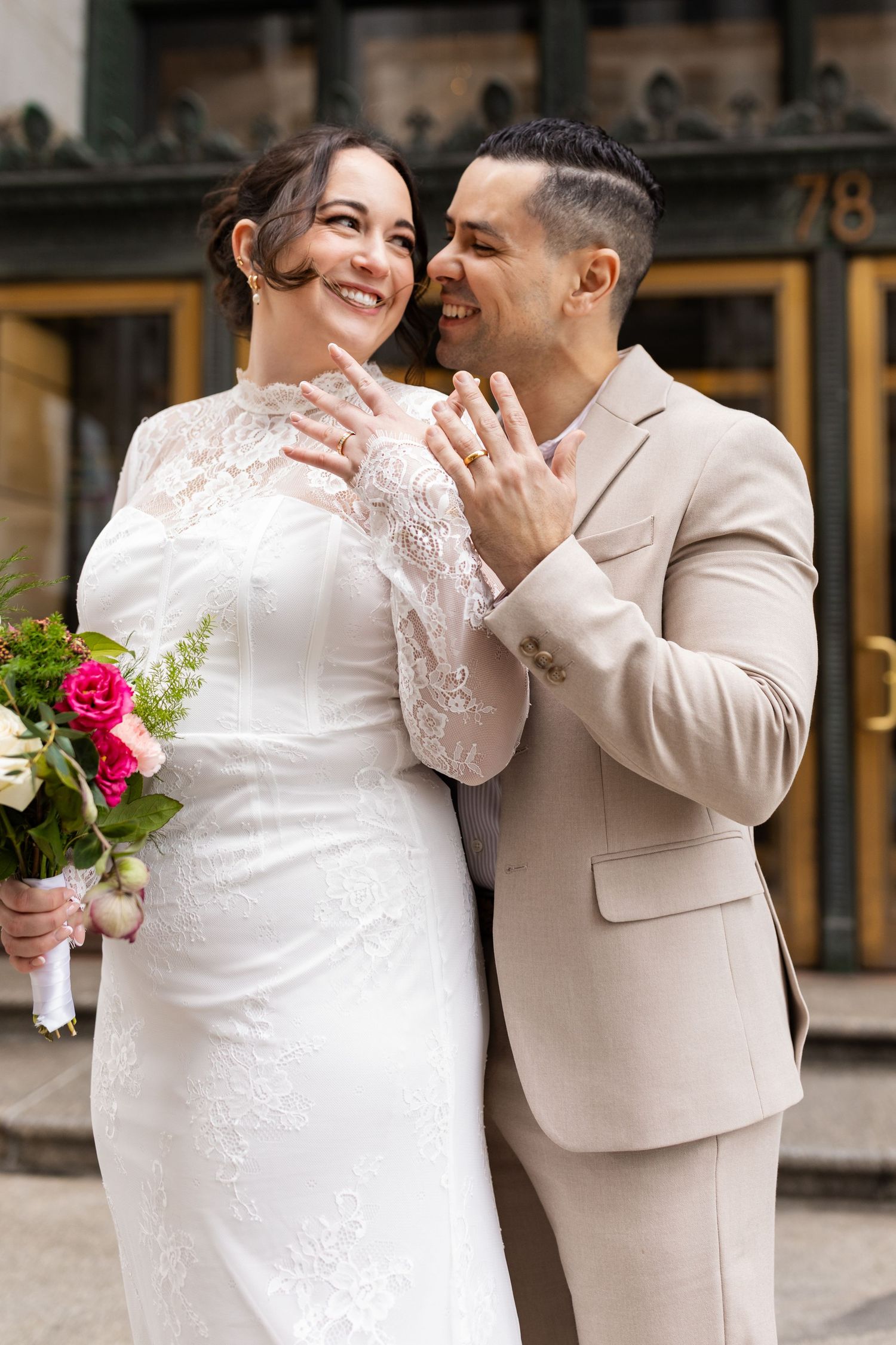 Joyful bride and groom sharing a tender moment, showing wedding rings, bride in lace gown holding pink bouquet.