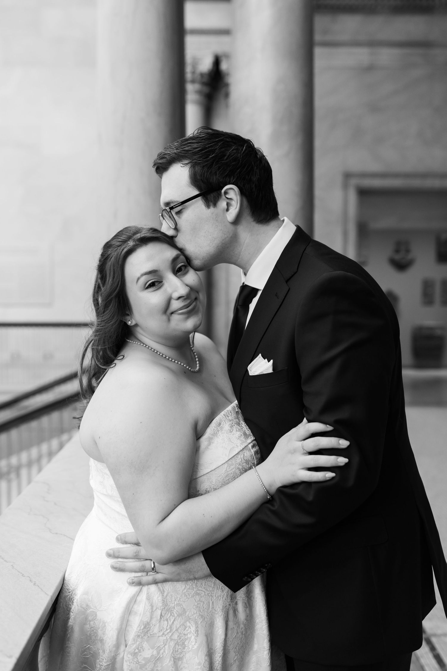 Black and white photo of groom kissing bride on cheek as she smiles at camera.