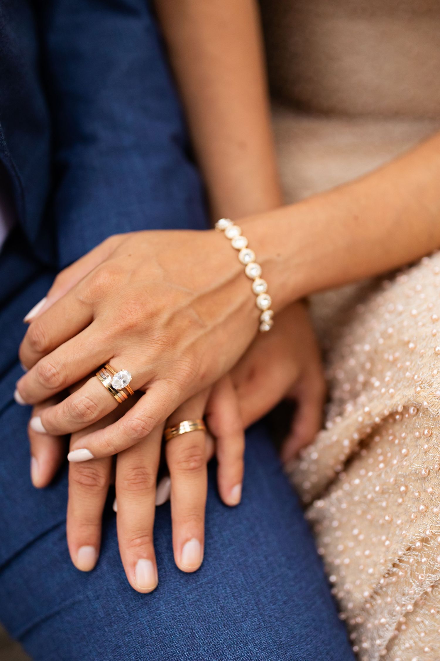 Close-up of couple's hands showing diamond engagement ring, gold wedding band, and pearl bracelet on blue suit.
