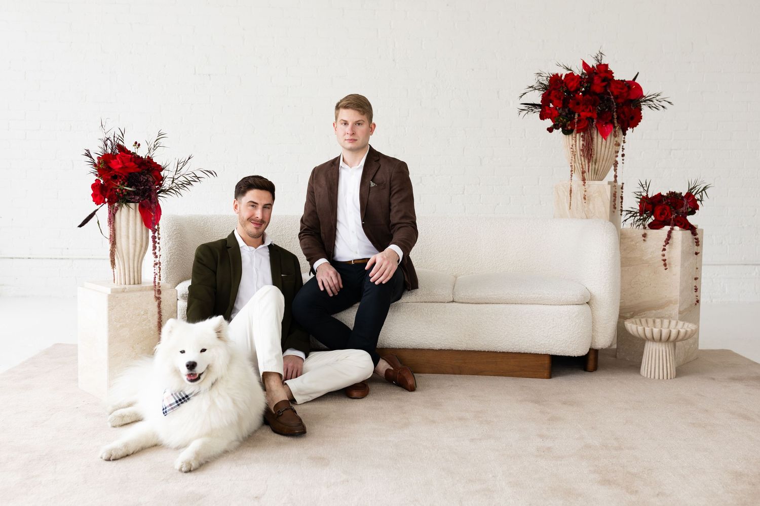 Two grooms smiling with their white Samoyed dog amid red floral arrangements in a studio.