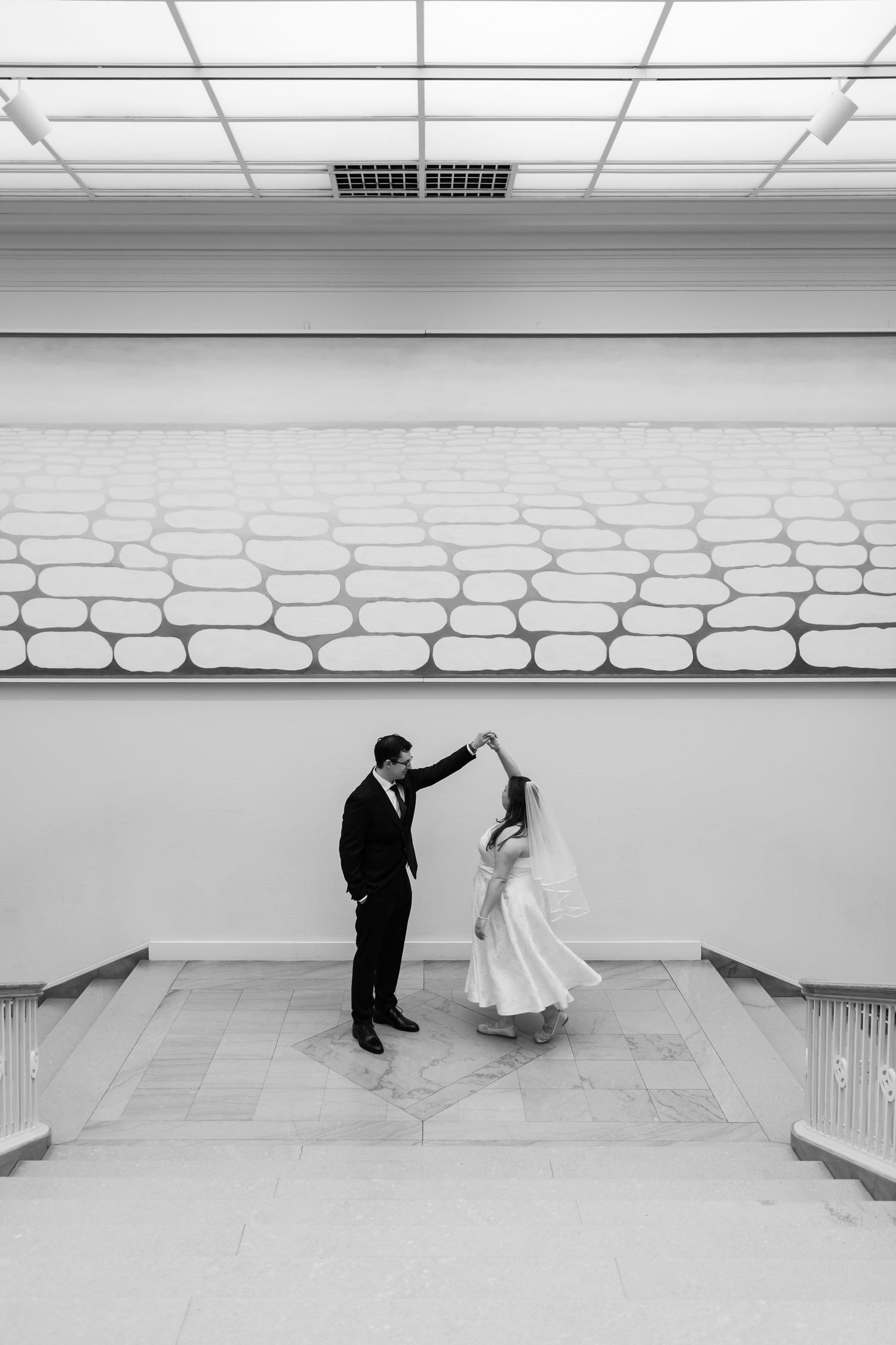 Black and white photo of bride and groom dancing elegantly in a bright modern museum interior.