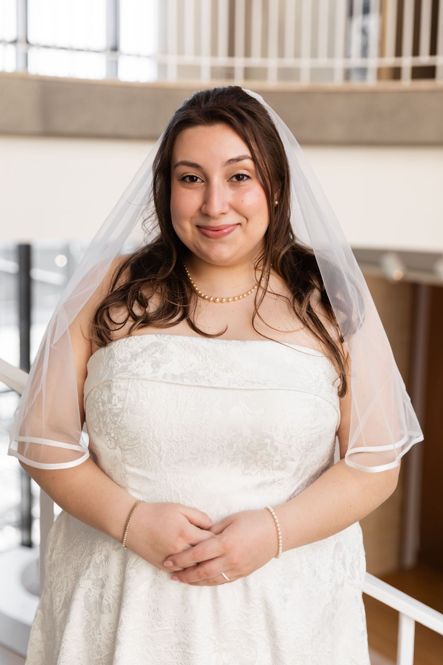 Smiling bride in white lace strapless gown, veil, and pearl necklace posing on staircase indoors.