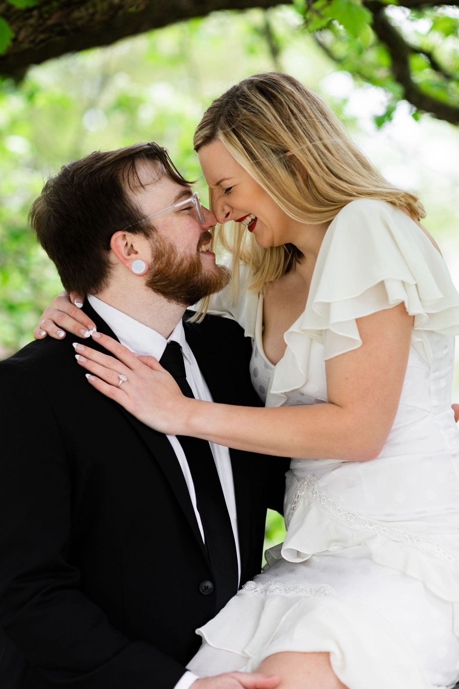 Laughing bride and groom share a joyful almost-kiss moment beneath a canopy of green trees.