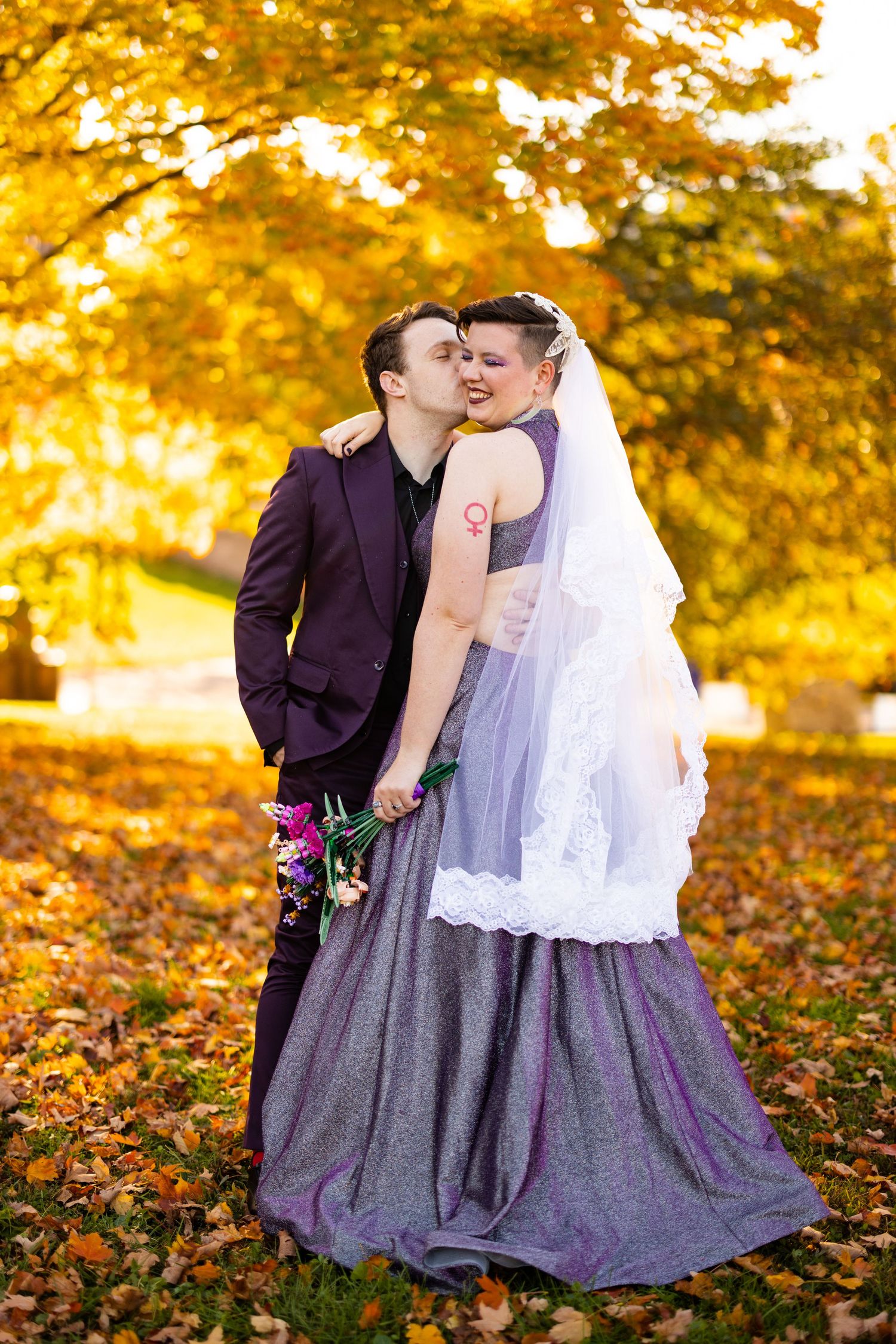 Groom kisses bride wearing purple gown and veil among golden autumn leaves in a park setting.