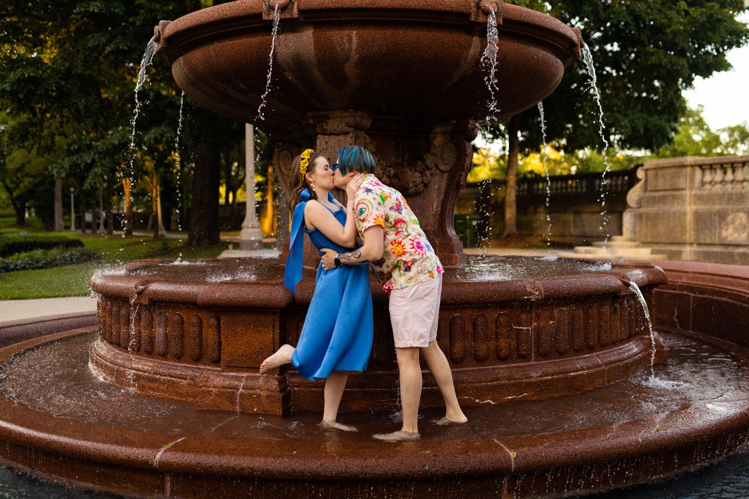 A couple shares a romantic kiss beside an ornate stone fountain in a lush green park.