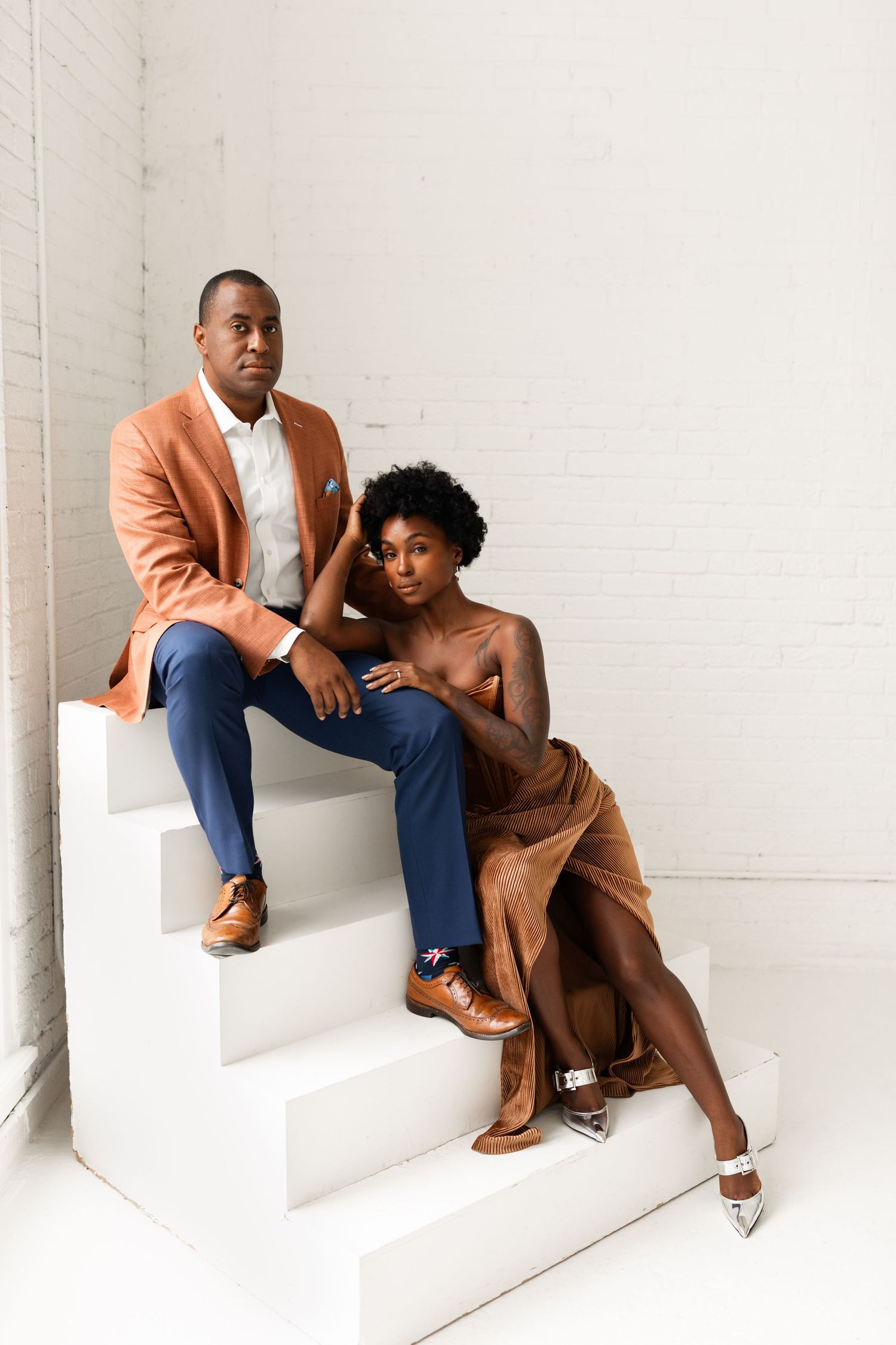 Elegant couple posing on white stairs, man in orange blazer and blue pants, woman in gold velvet dress.