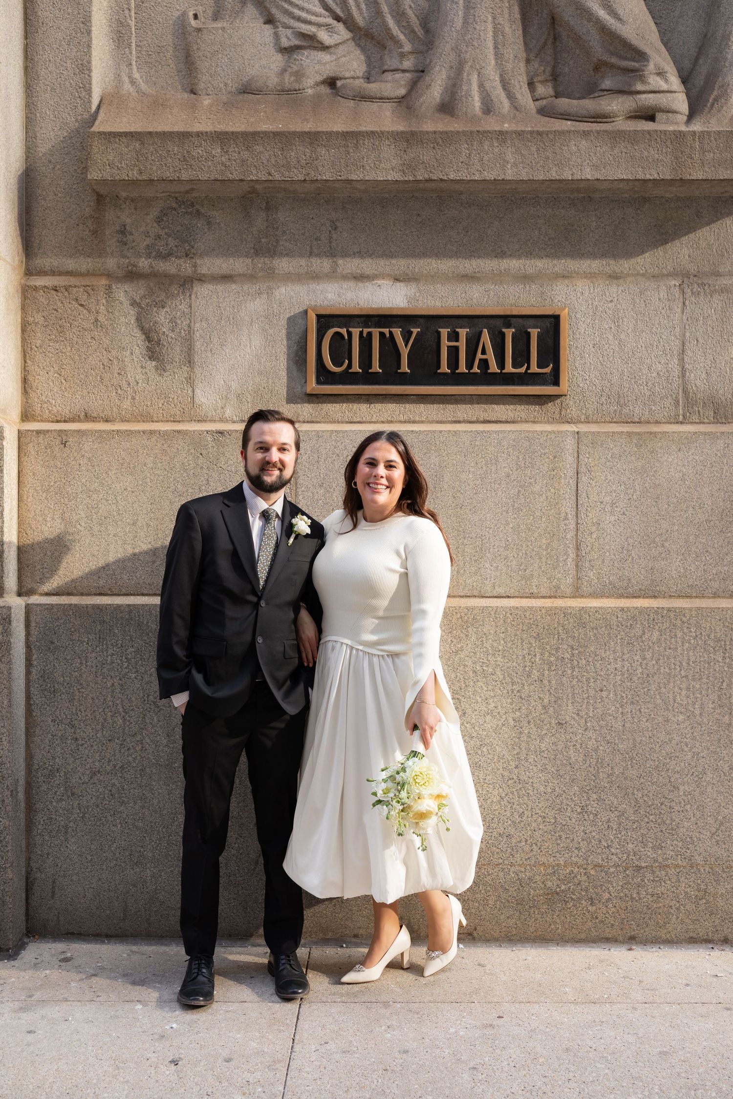 Newlyweds pose smiling in front of a City Hall sign, bride holding a white and yellow bouquet.