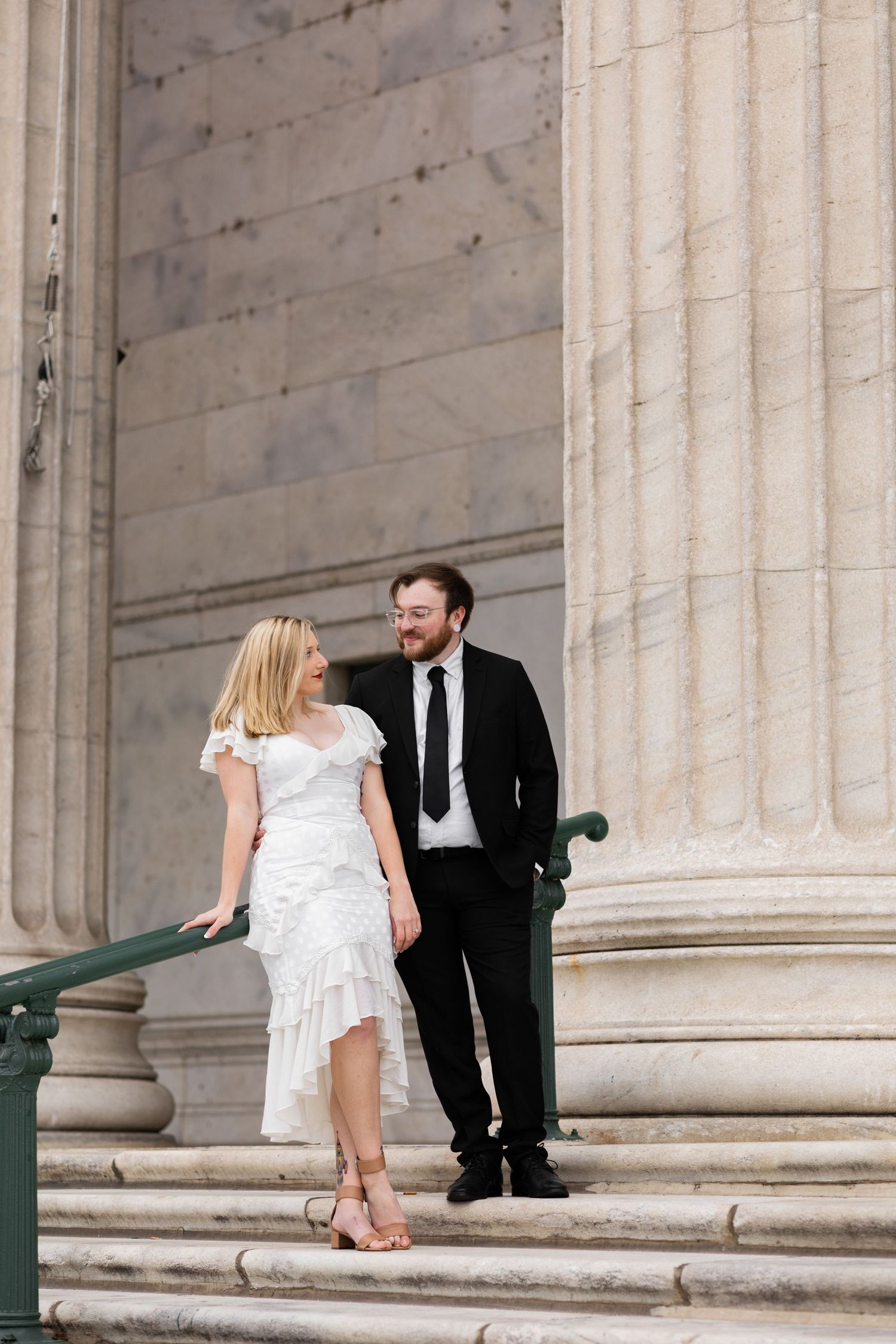 Bride and groom share a glance while posing on steps beside towering stone columns of a landmark.