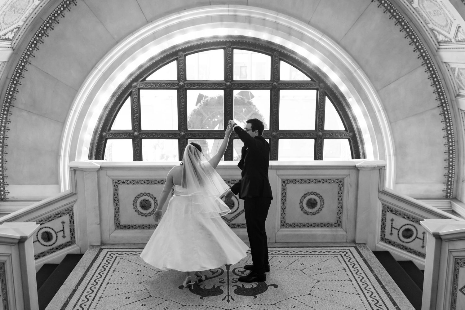 Black and white photo of bride and groom dancing beneath an arched window in ornate venue.