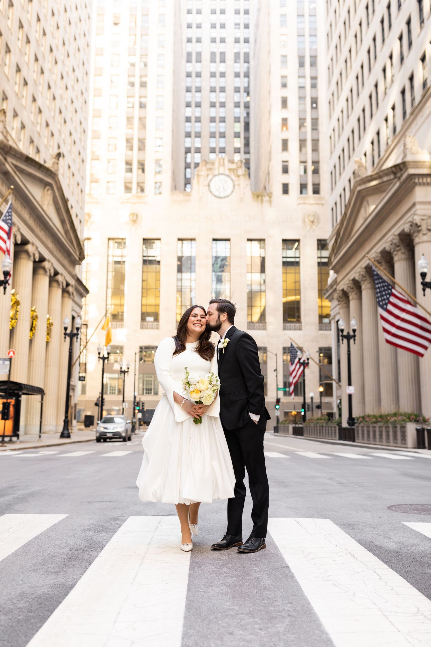 Groom kisses bride on the cheek as she holds a yellow bouquet on a crosswalk in downtown Chicago.