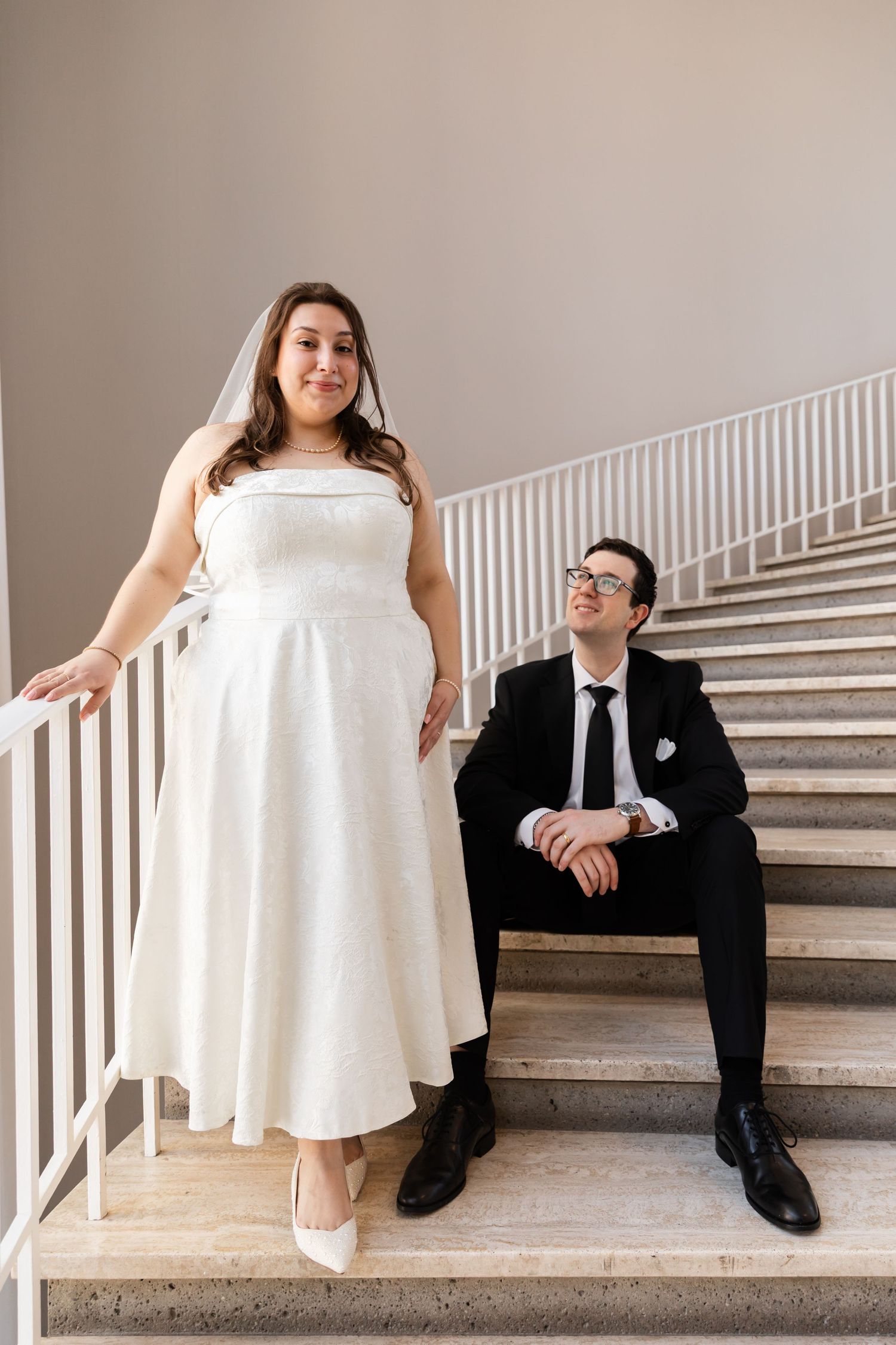 Bride standing on marble stairs while groom sits looking up at her adoringly in elegant venue.