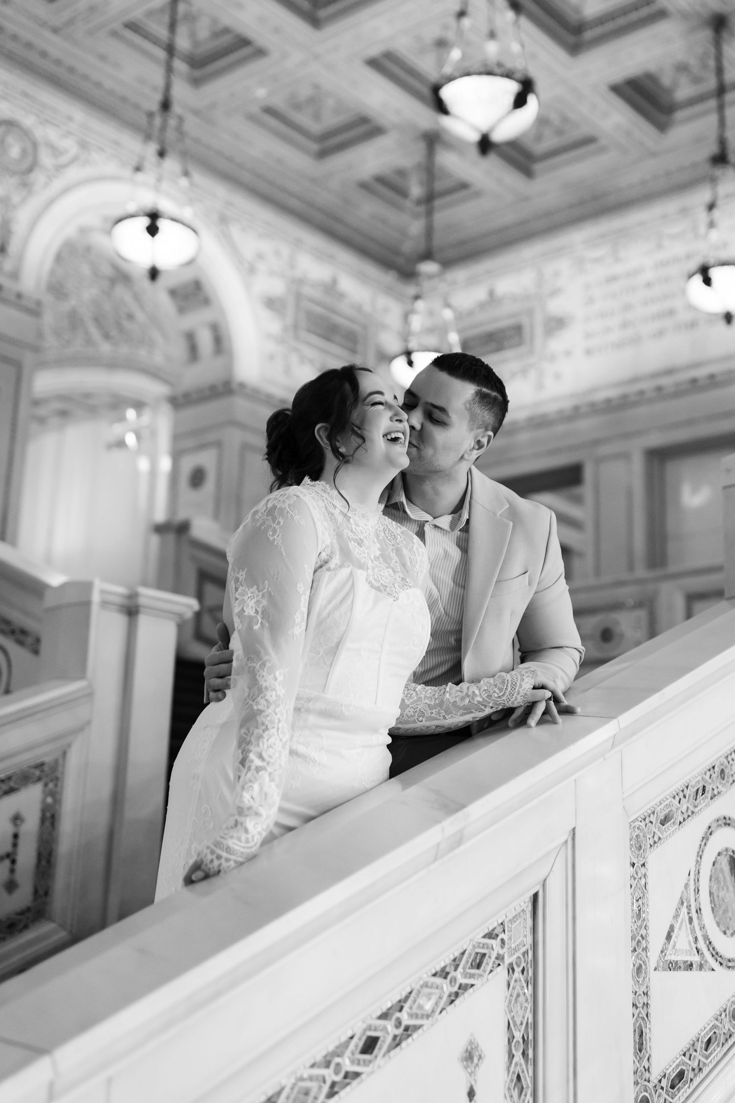 Black and white photo of a laughing bride and groom leaning on a marble staircase railing.