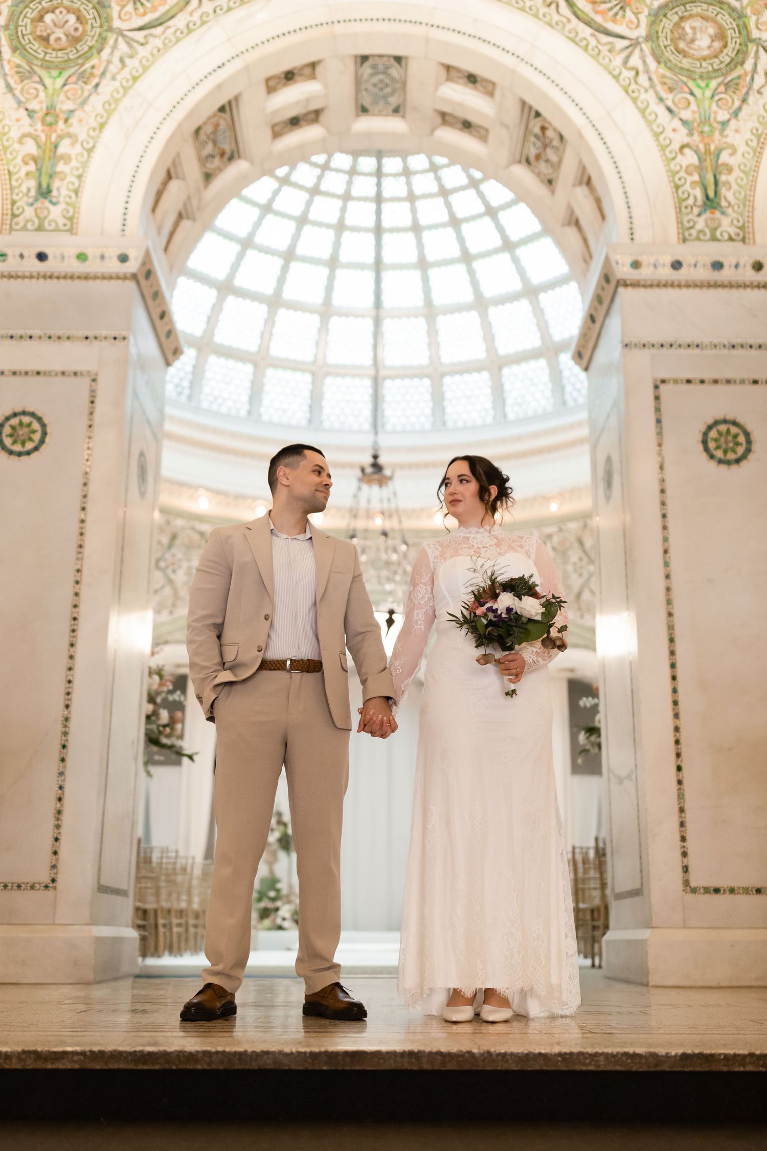 Bride and groom holding hands under a stunning domed glass ceiling in an elegant historic building.