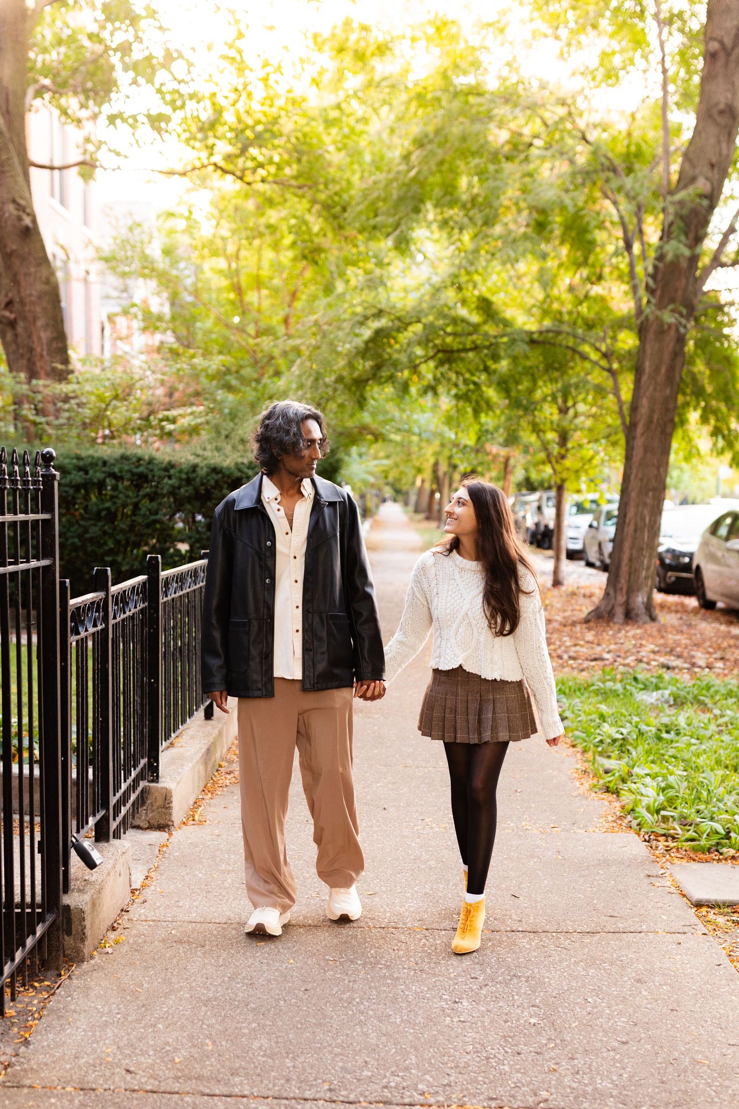 Interracial couple walking hand in hand on tree-lined urban sidewalk in autumn, smiling at each other.