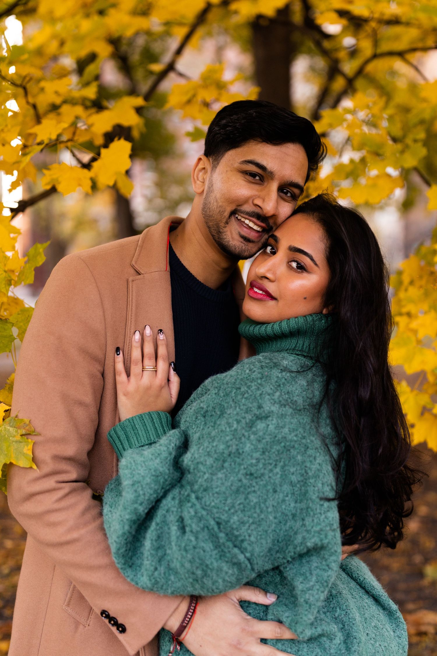 Couple embraces warmly in front of golden autumn leaves, woman in teal sweater and man in beige coat.