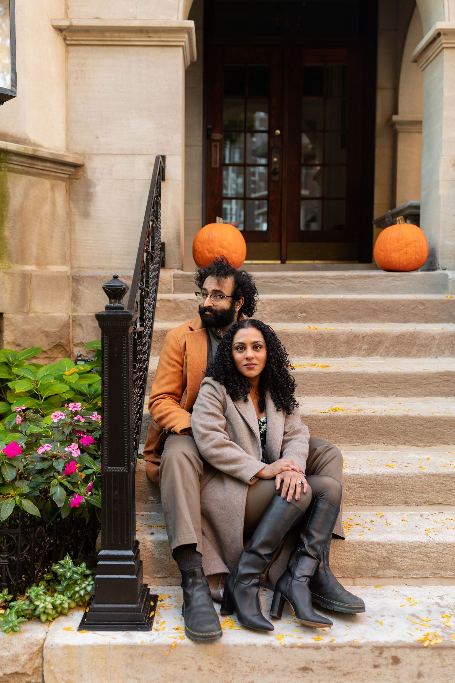 A couple sits together on stone steps decorated with pumpkins outside a classic building in fall.