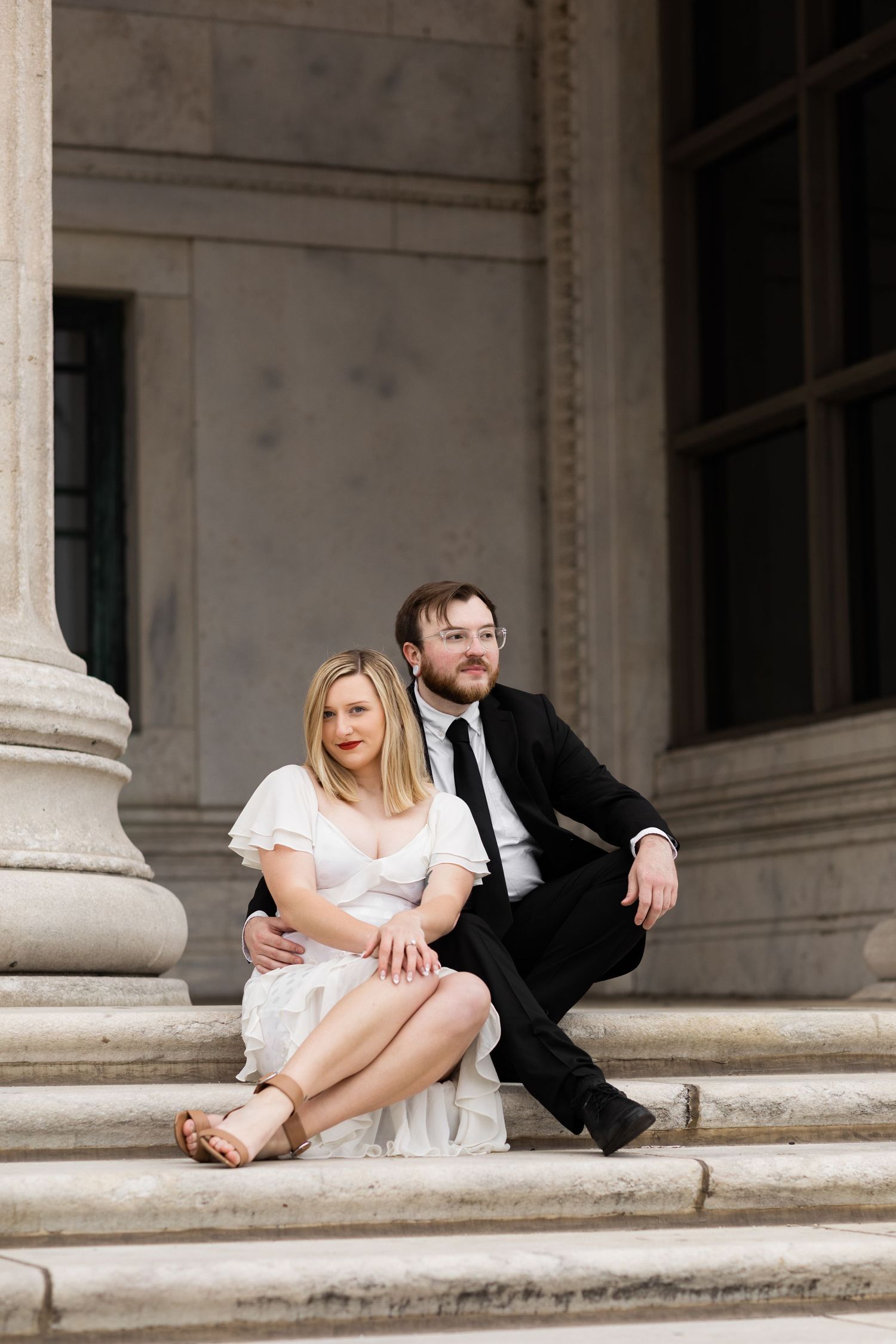 Bride and groom sit together on marble steps of a grand stone building with tall columns.