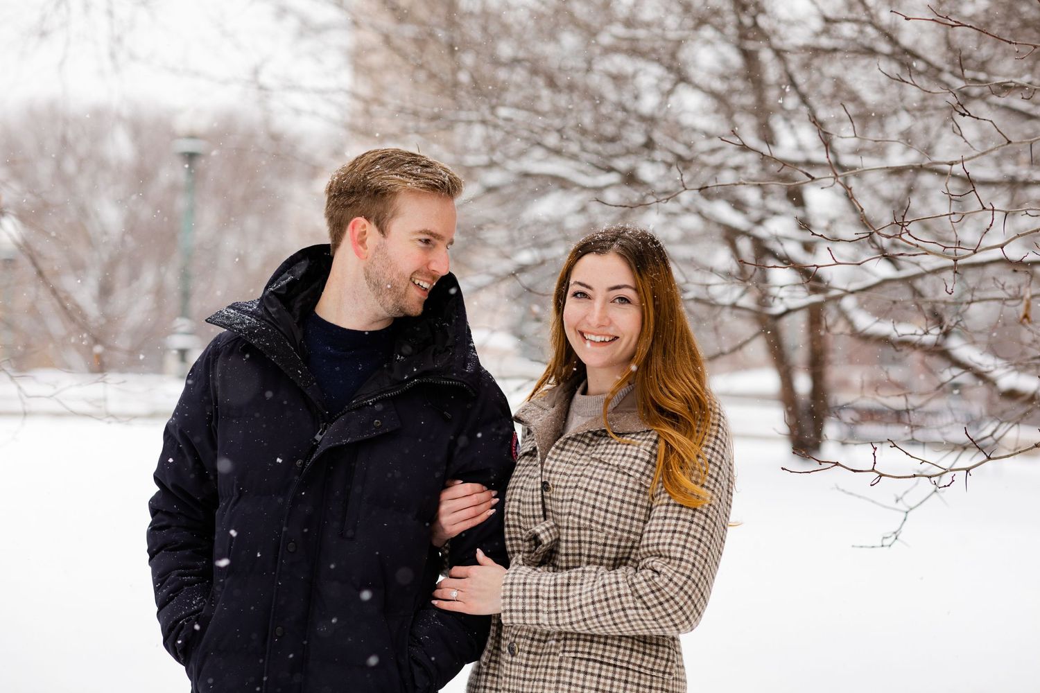 Smiling couple embracing in a snowy winter park during a light snowfall.