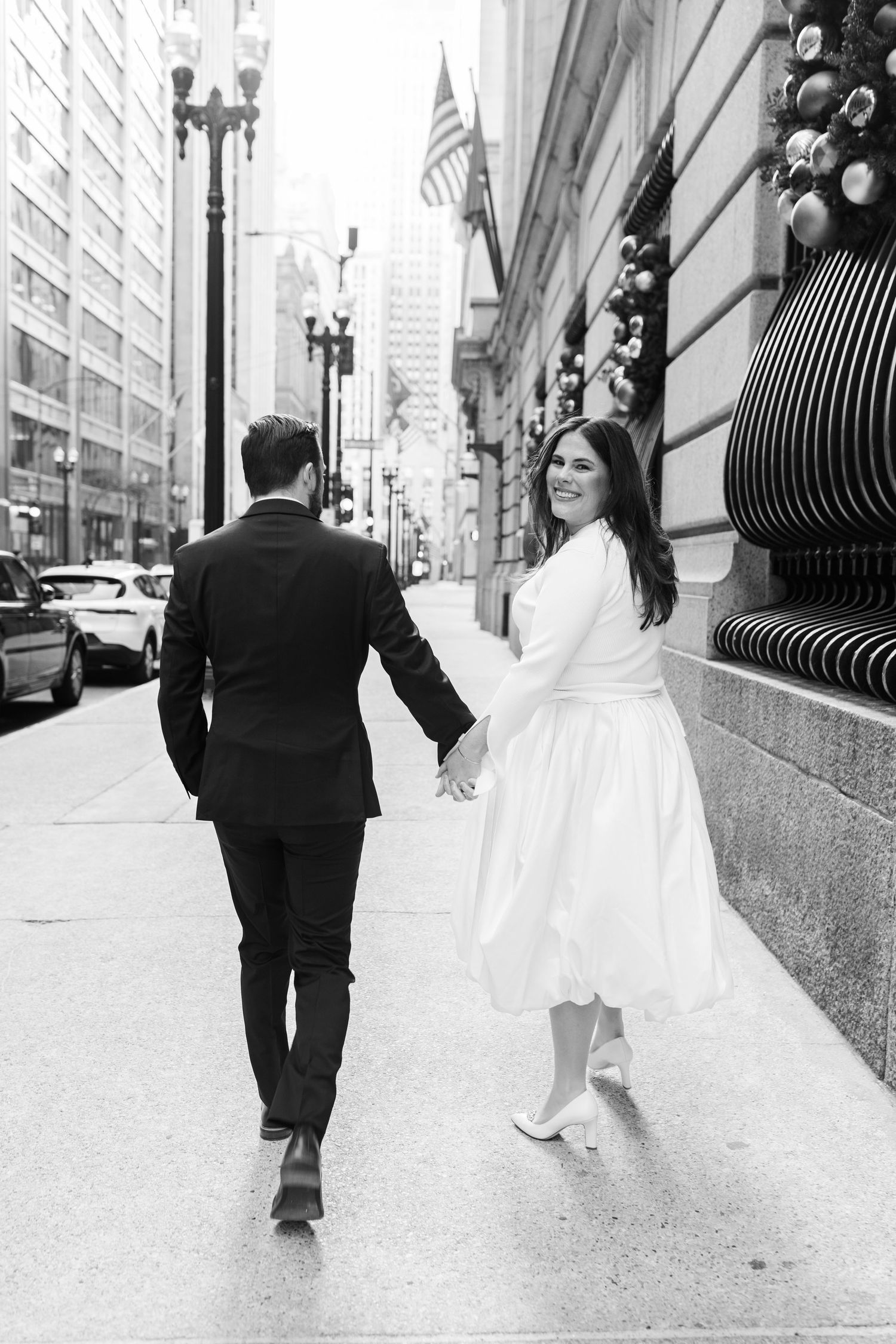 Bride in white dress and groom in black suit walk hand-in-hand down a city sidewalk, black and white photo.