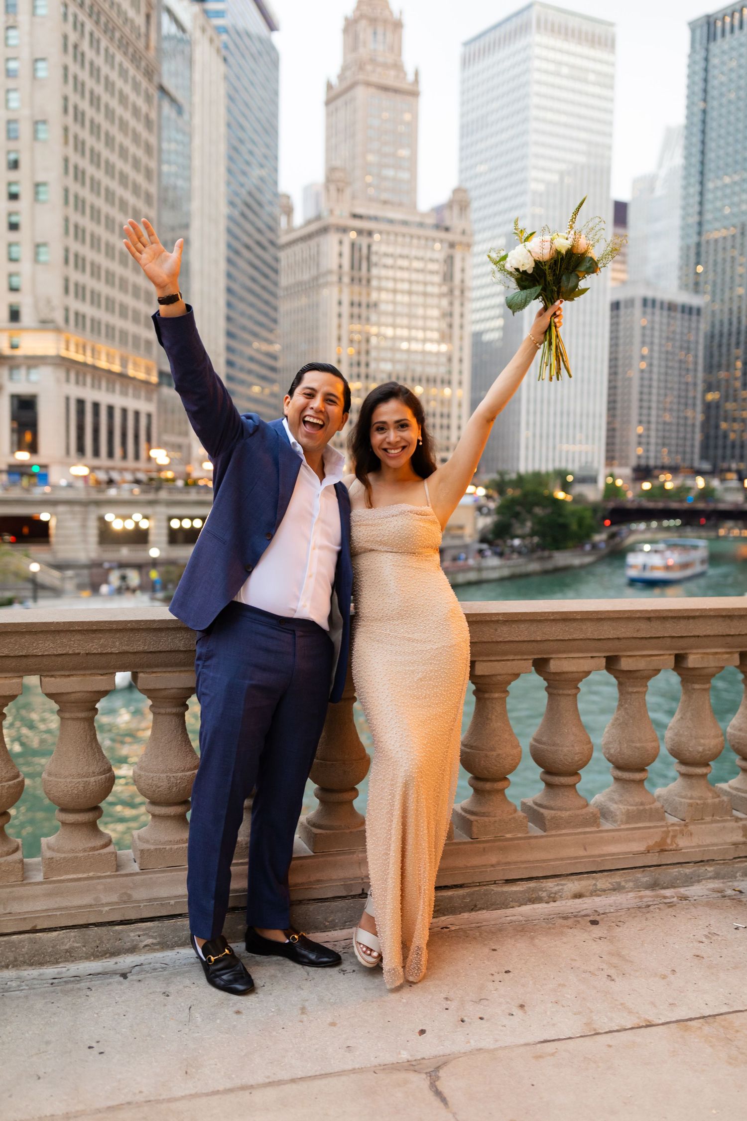 Joyful couple celebrating with arms raised on Chicago Riverwalk balustrade with city skyline at dusk.