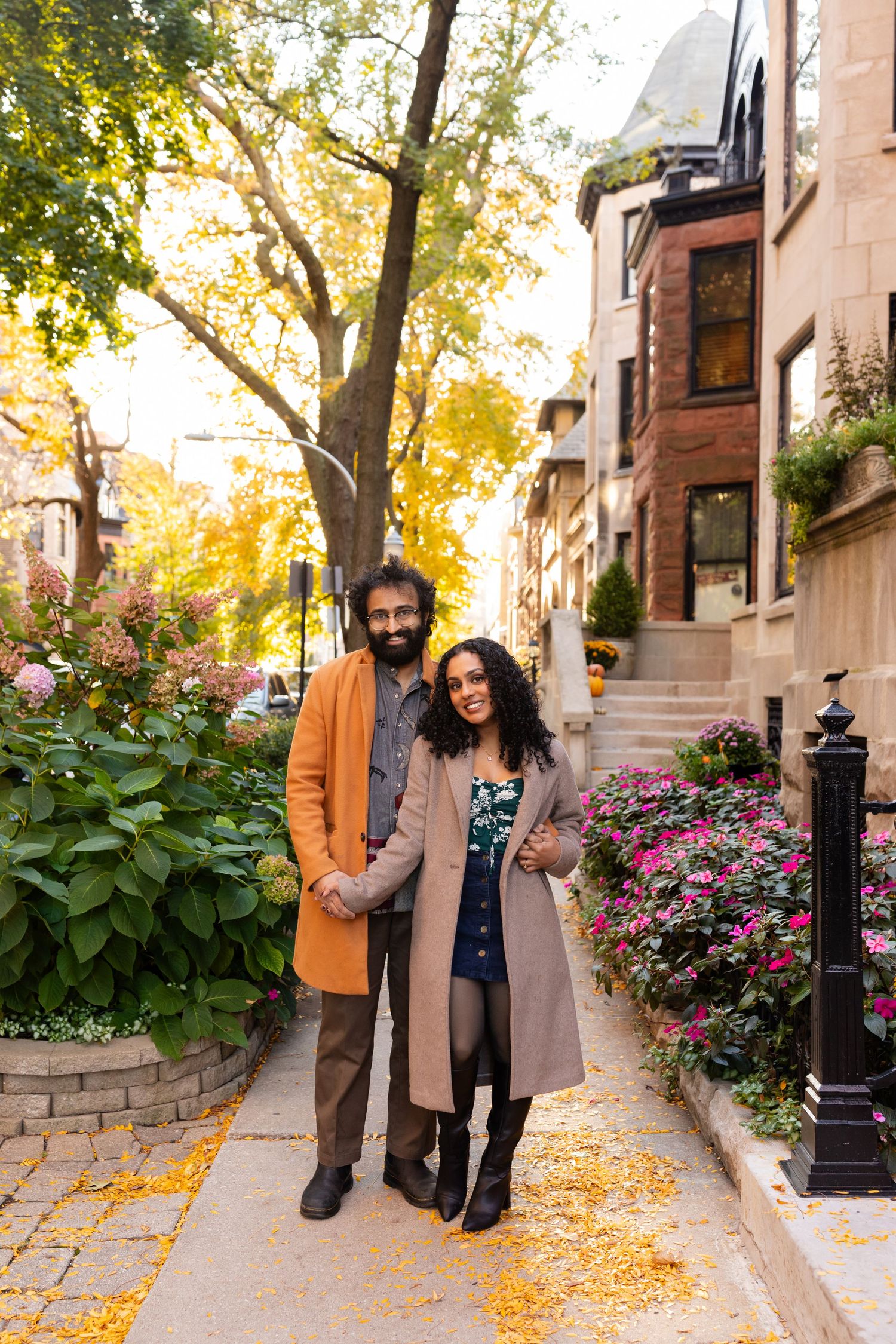 Stylish couple posing on a flower-lined sidewalk during a golden autumn day.
